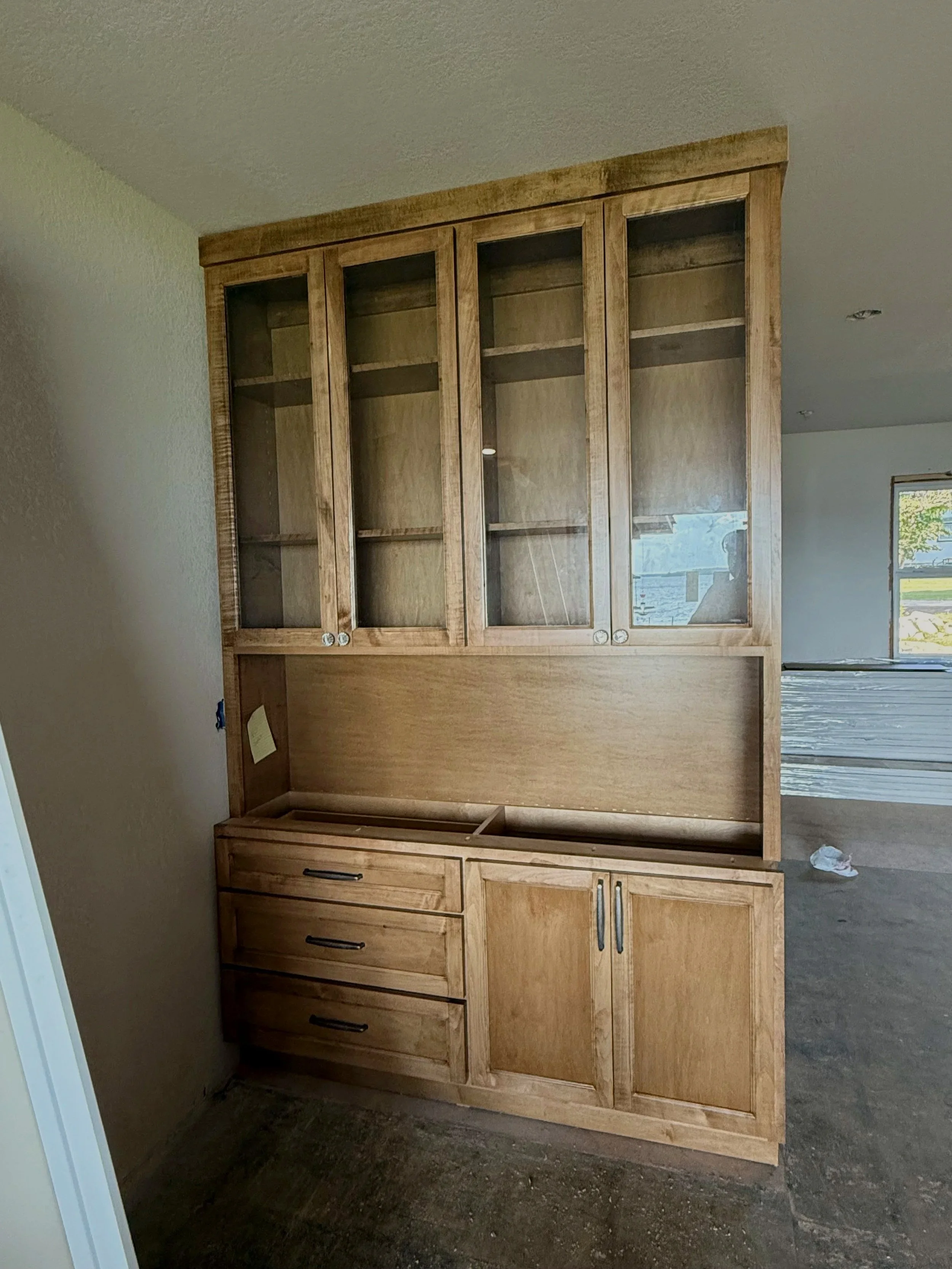 A large, wooden kitchen cabinet with glass doors on upper sections and solid doors on lower sections, against a beige wall in a room with a window.