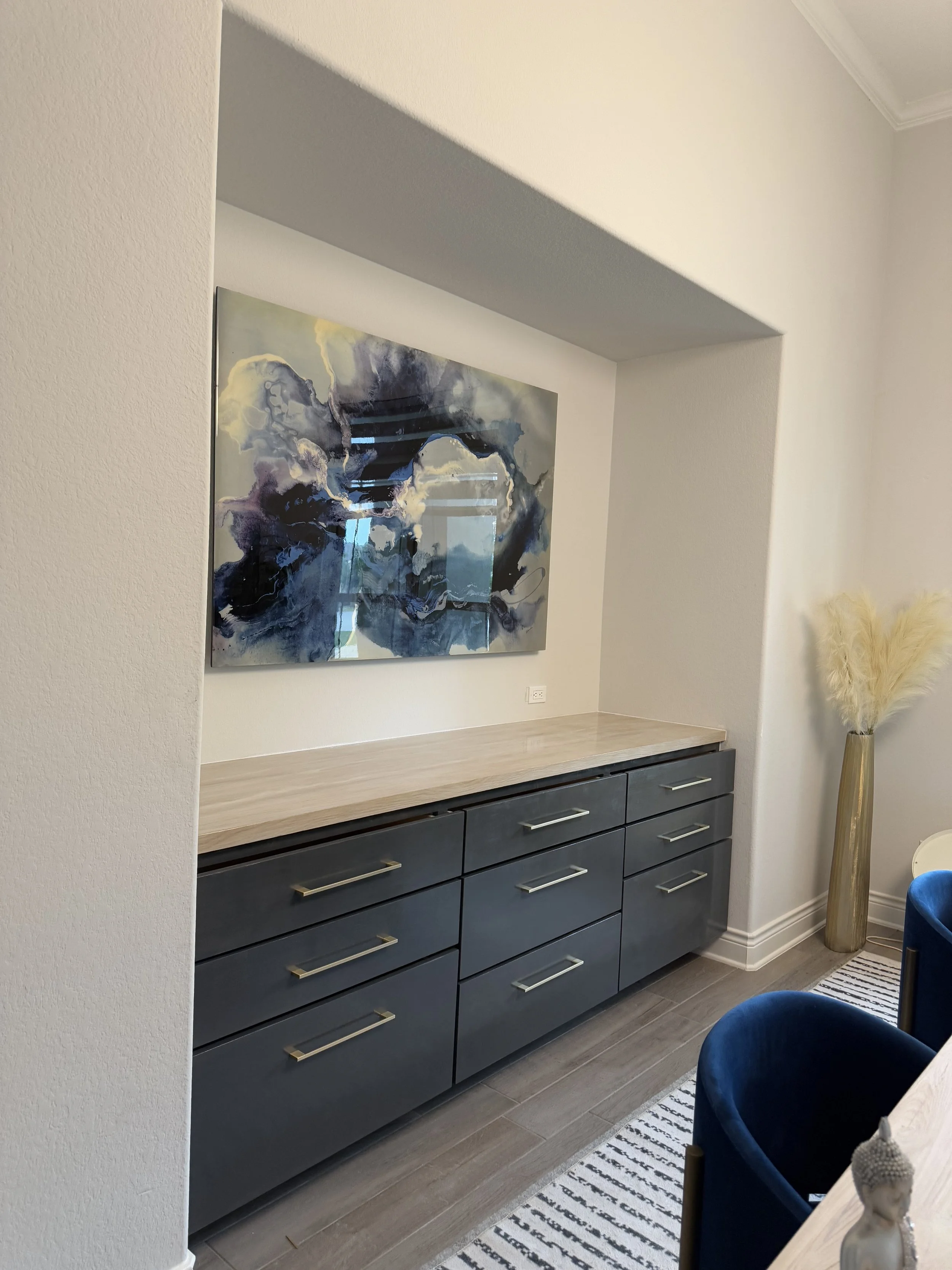 A modern sideboard with multiple drawers in navy blue, with brass handles. Above it on the wall, there is an abstract painting with shades of blue, white, gray, and yellow. To the right, a tall decorating vase with beige pampas grass inside. Part of 