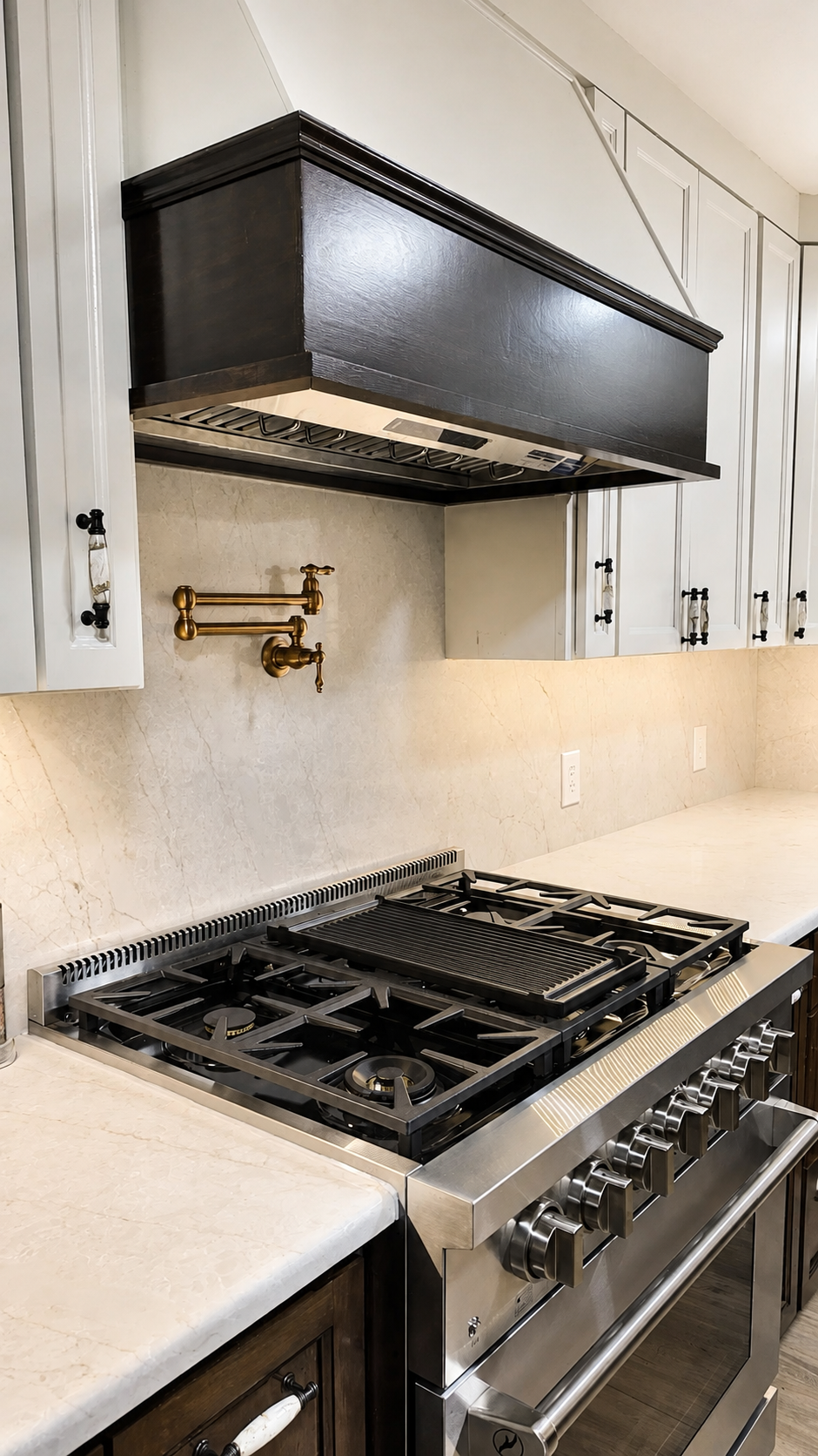 Modern kitchen with white cabinetry, a beige marble countertop, and a stainless steel gas stove. There are black and copper accents, including a black range hood and copper pipes underneath the cabinets. Electrical outlets are visible on the backsplash, and a window is on the right side.