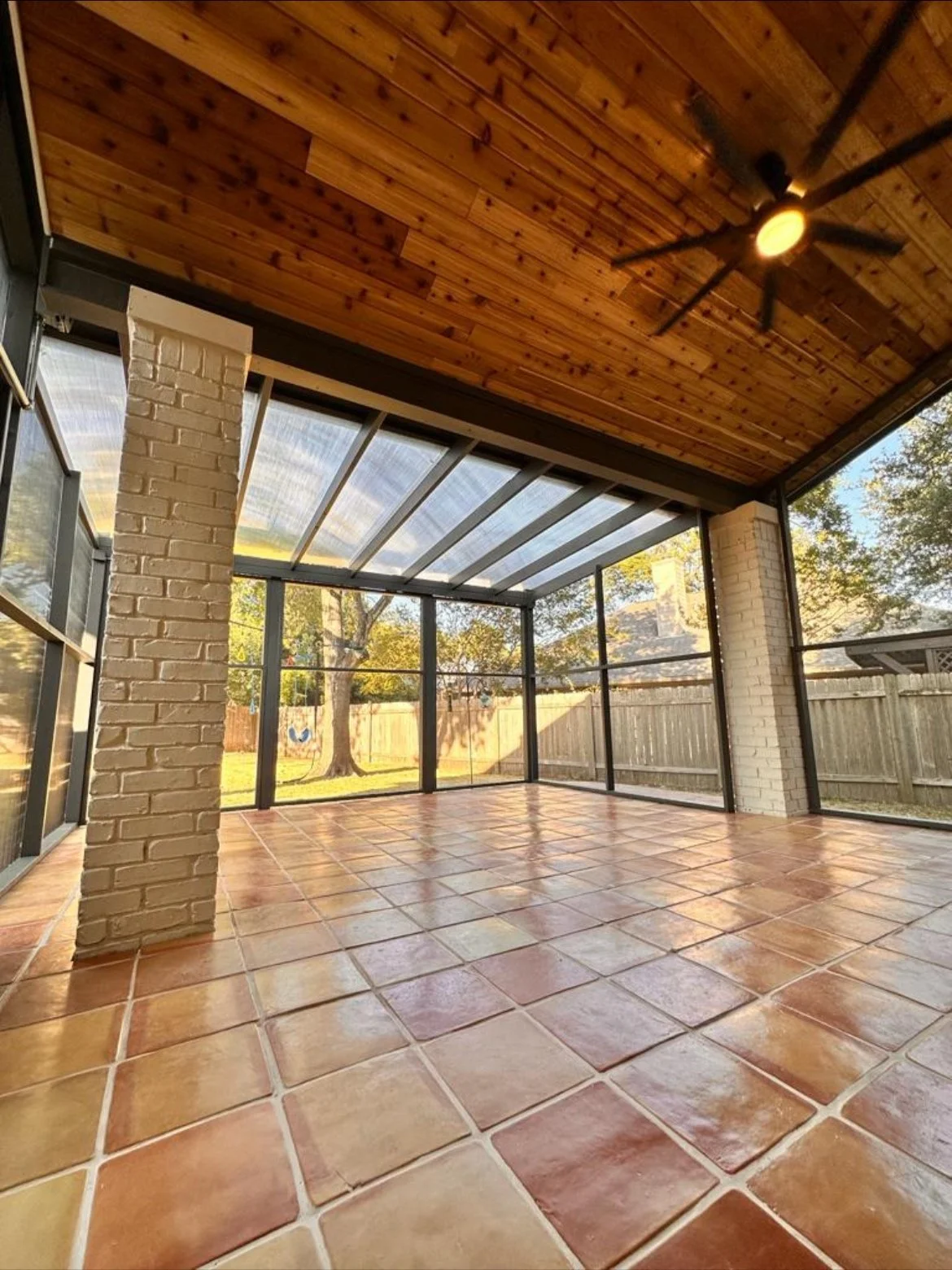 Enclosed patio with terracotta tile flooring, brick columns, wooden ceiling, ceiling fan, and large screen windows showing backyard with trees and wooden fence.