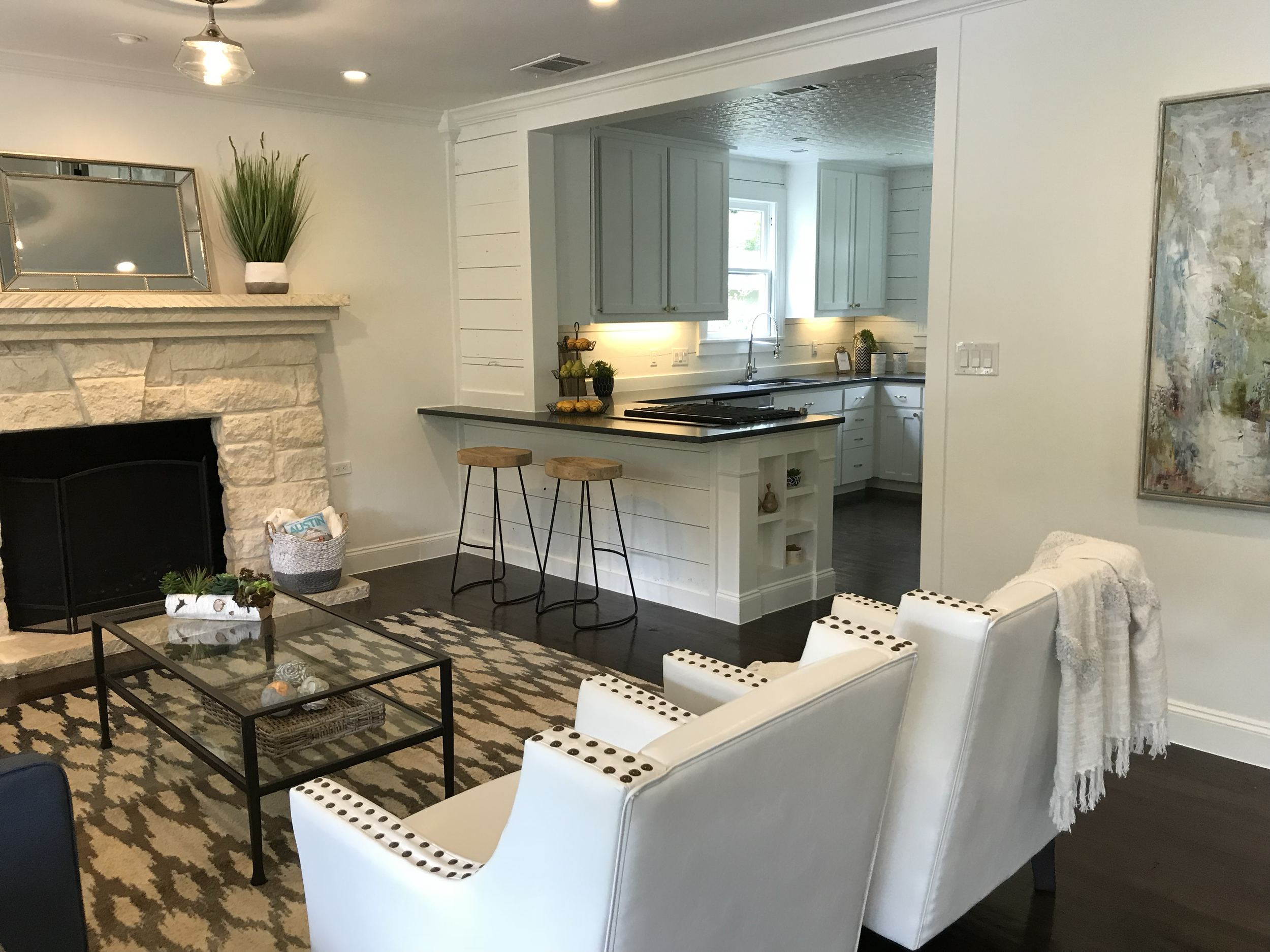A living room and kitchen area with white walls and dark hardwood floors. The living room has a stone fireplace with a mirror and a tall plant on the mantel, a patterned rug, and white armchairs with black polka dots. The kitchen has light blue cabinets, black countertops, a window above the sink, and small decorative items on the counter. There are also two wooden barstools at the kitchen counter.