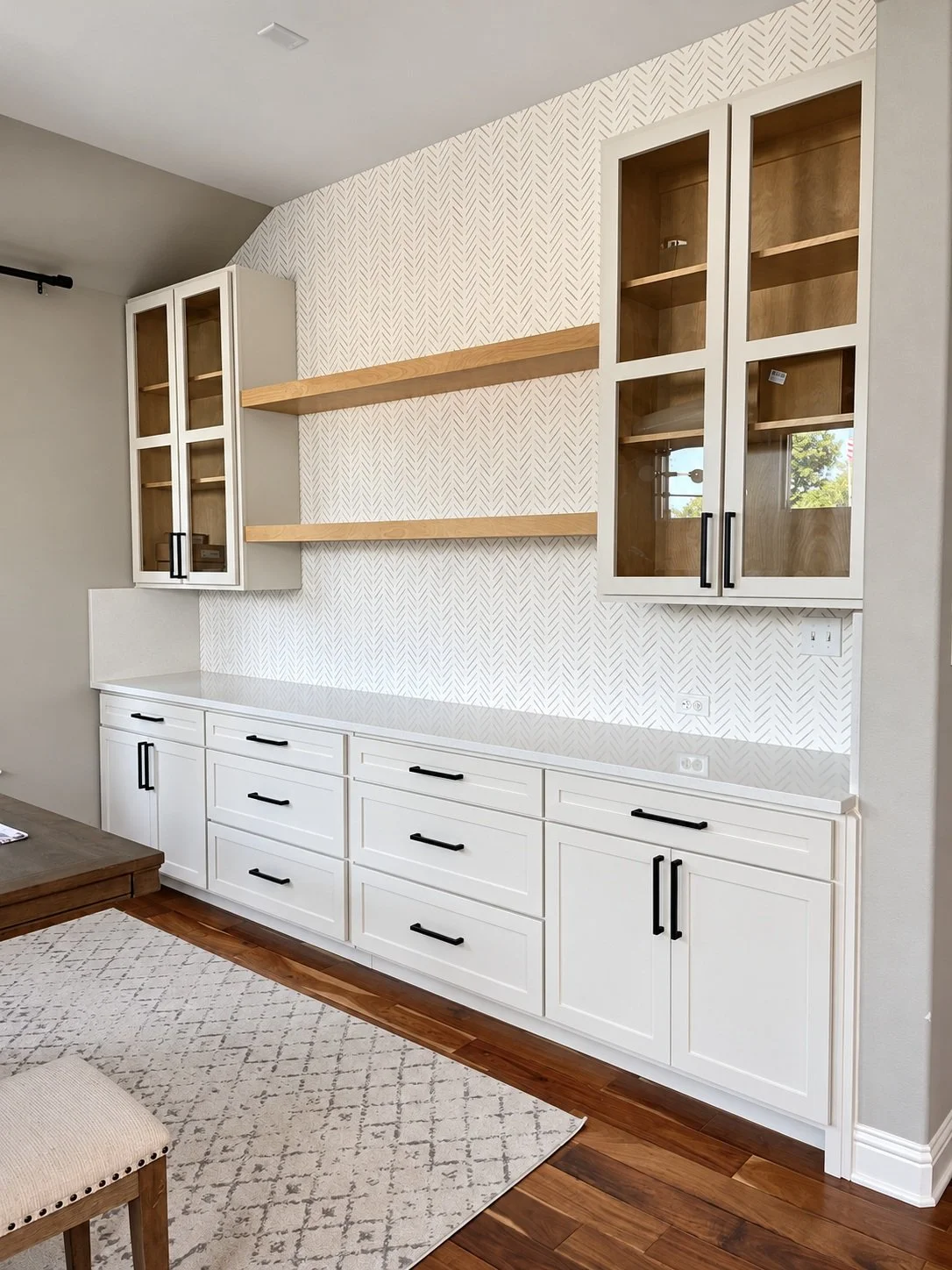 White built-in cabinetry with glass-front upper doors, warm wood interiors, floating shelves, and black hardware against a patterned wall.