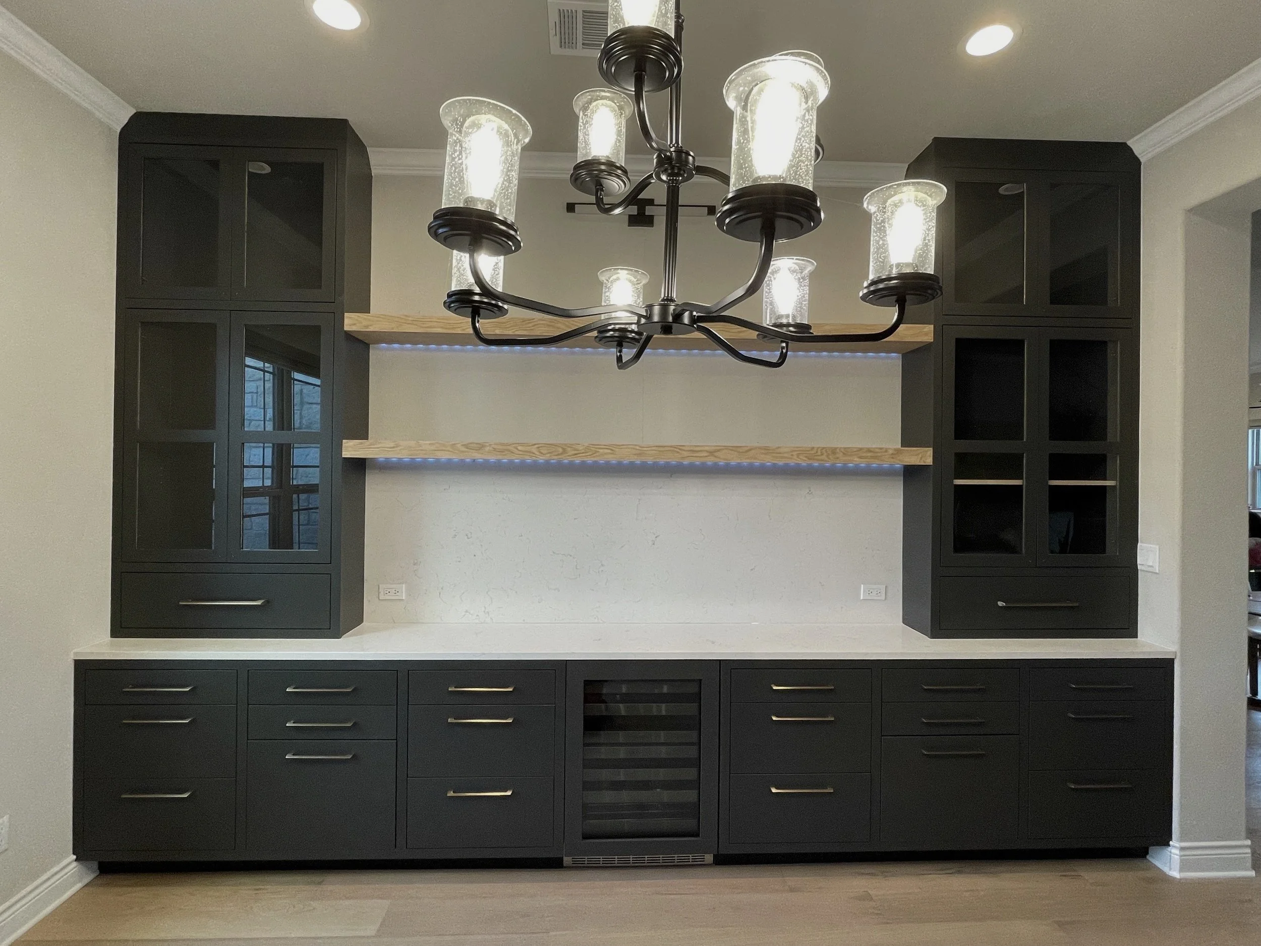 Modern black kitchen cabinets with glass doors, a built-in wine cooler, and a wooden shelf, with a chandelier hanging from the ceiling.