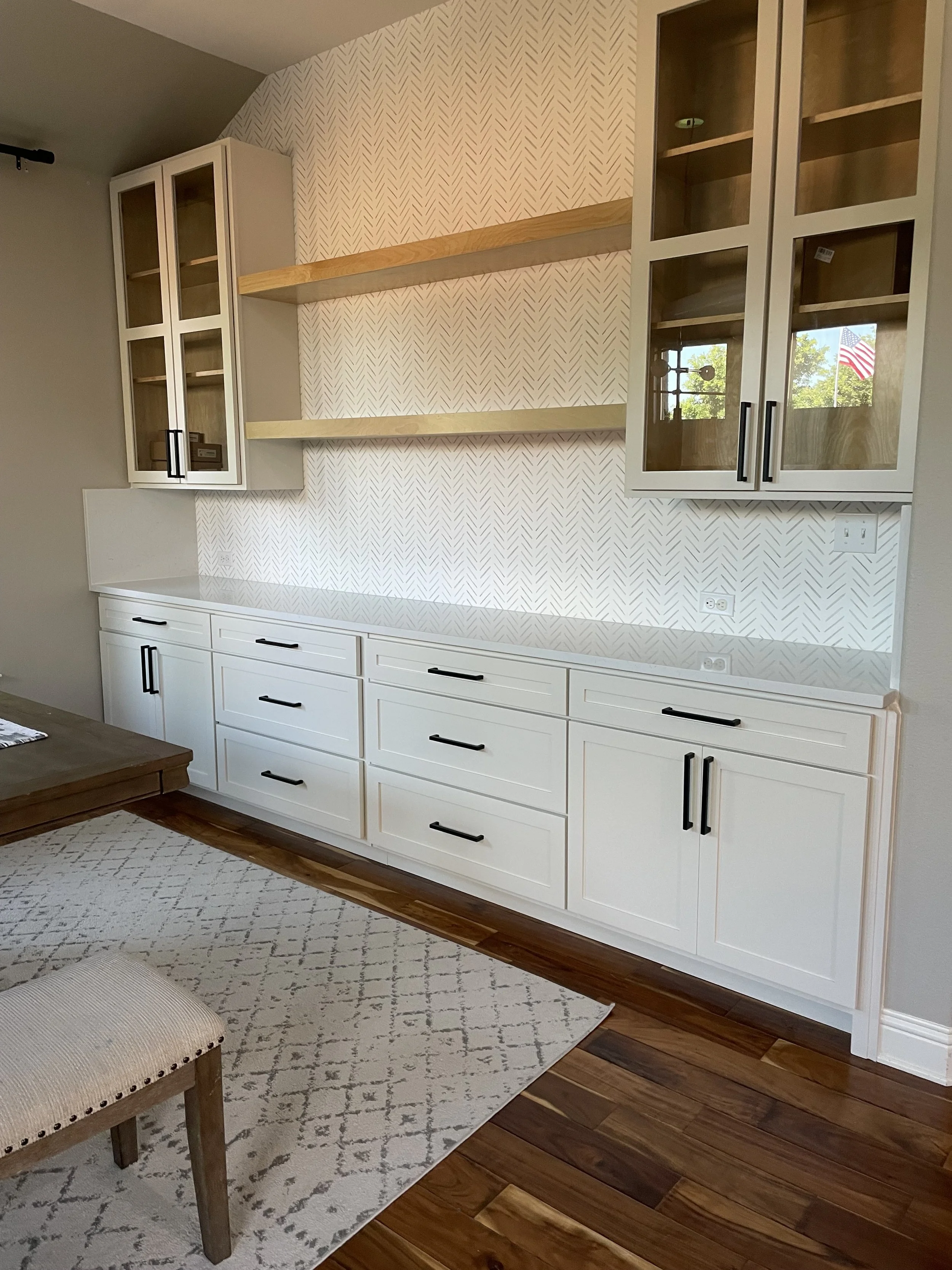 Built-in white cabinetry with black handles, featuring glass-front doors, mounted on a wall with a patterned wallpaper. Below the cabinets, there is a white countertop and drawers. Part of a rug and a wooden bench with beige upholstery are visible in