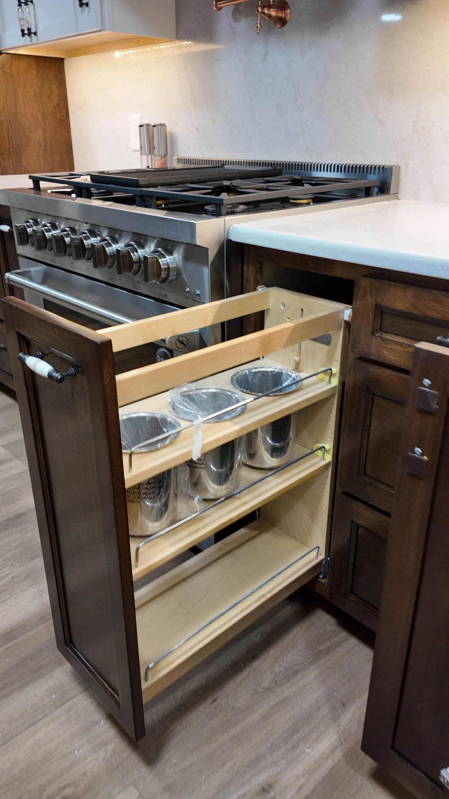 Kitchen with a stainless steel oven and stove, partially opened drawer with multiple metal trash bins, wooden and dark brown cabinets, and a light-colored countertop.