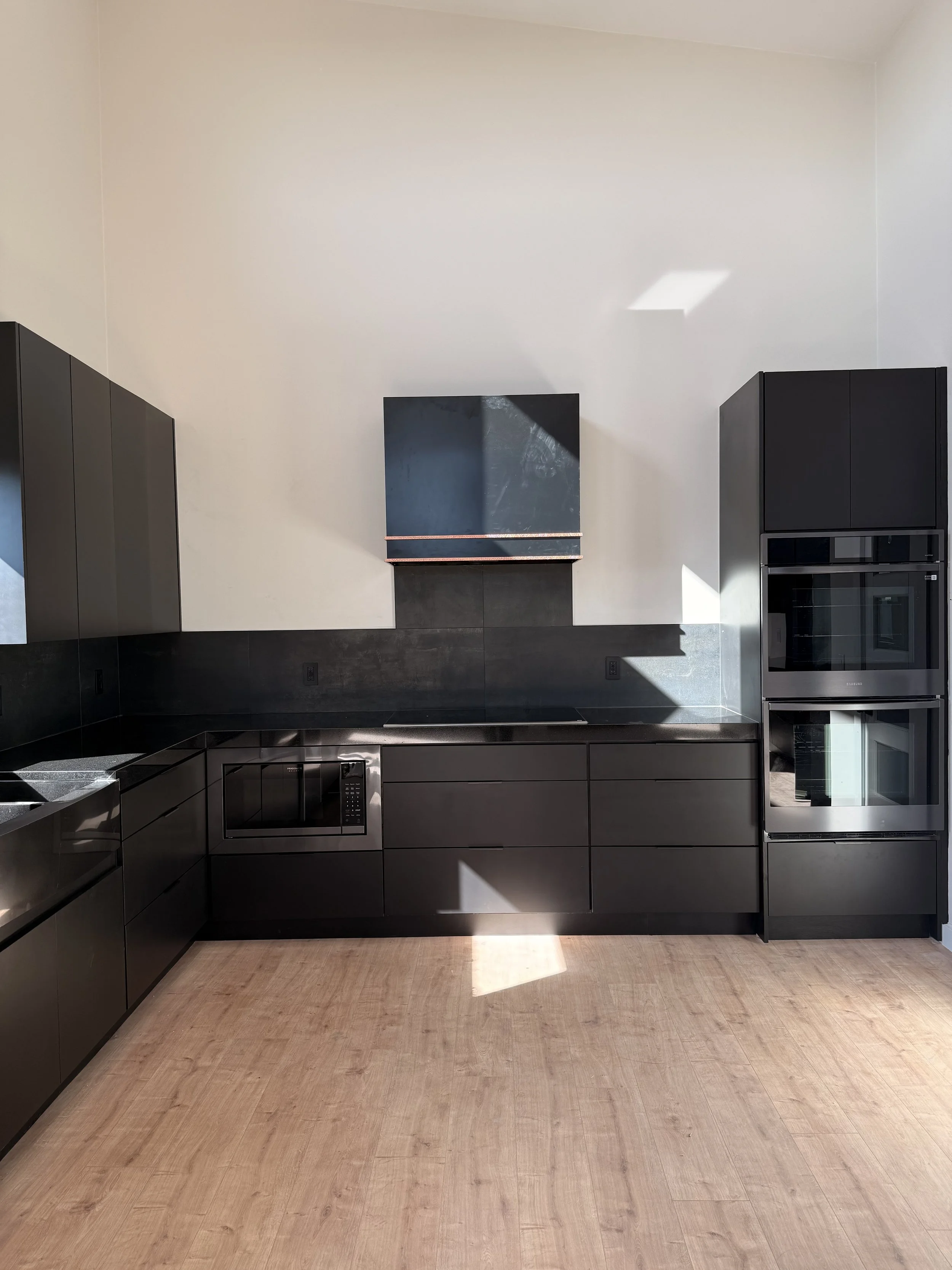 A modern kitchen with black cabinetry, stainless steel appliances, and light-colored hardwood flooring. There is a large, plain wall with sunlight casting shadows, and a black vent hood above the stove.