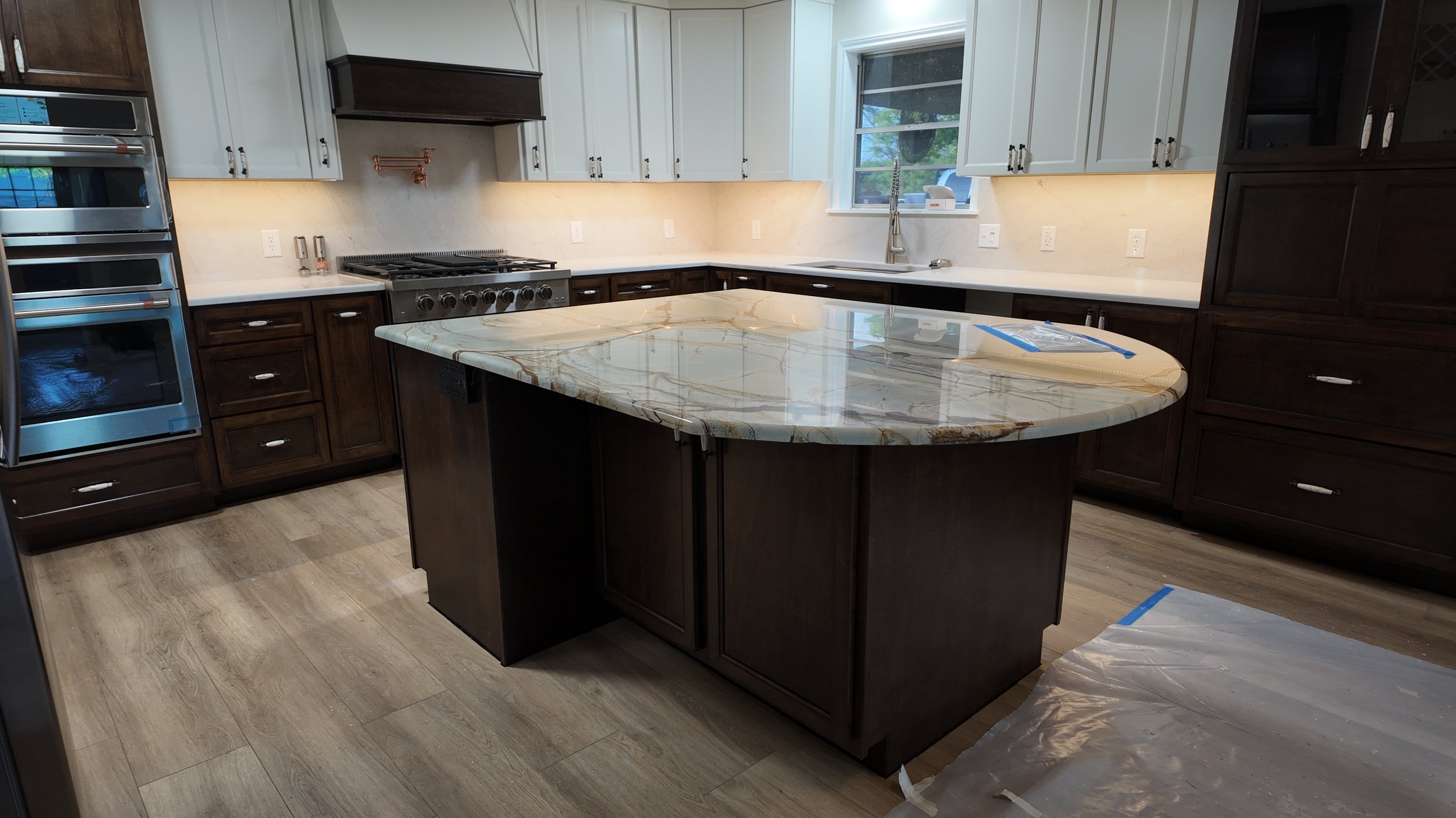 A modern kitchen with dark wood cabinets, white upper cabinets, a marble island, stainless steel appliances, and a window above the sink.