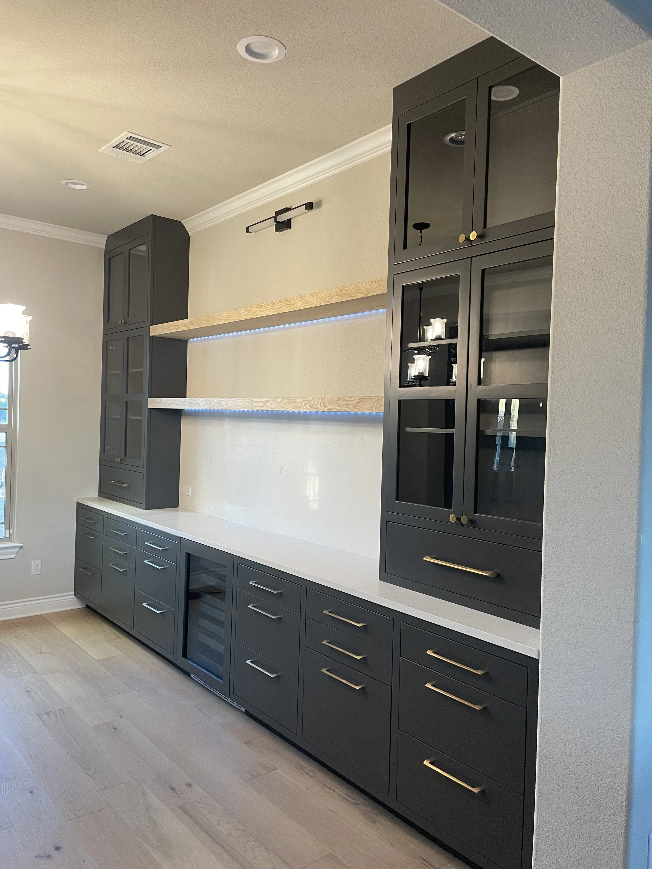 Modern kitchen cabinetry with dark gray cabinets, gold handles, glass-paneled doors, and LED lighting, with a white countertop and light wood flooring.