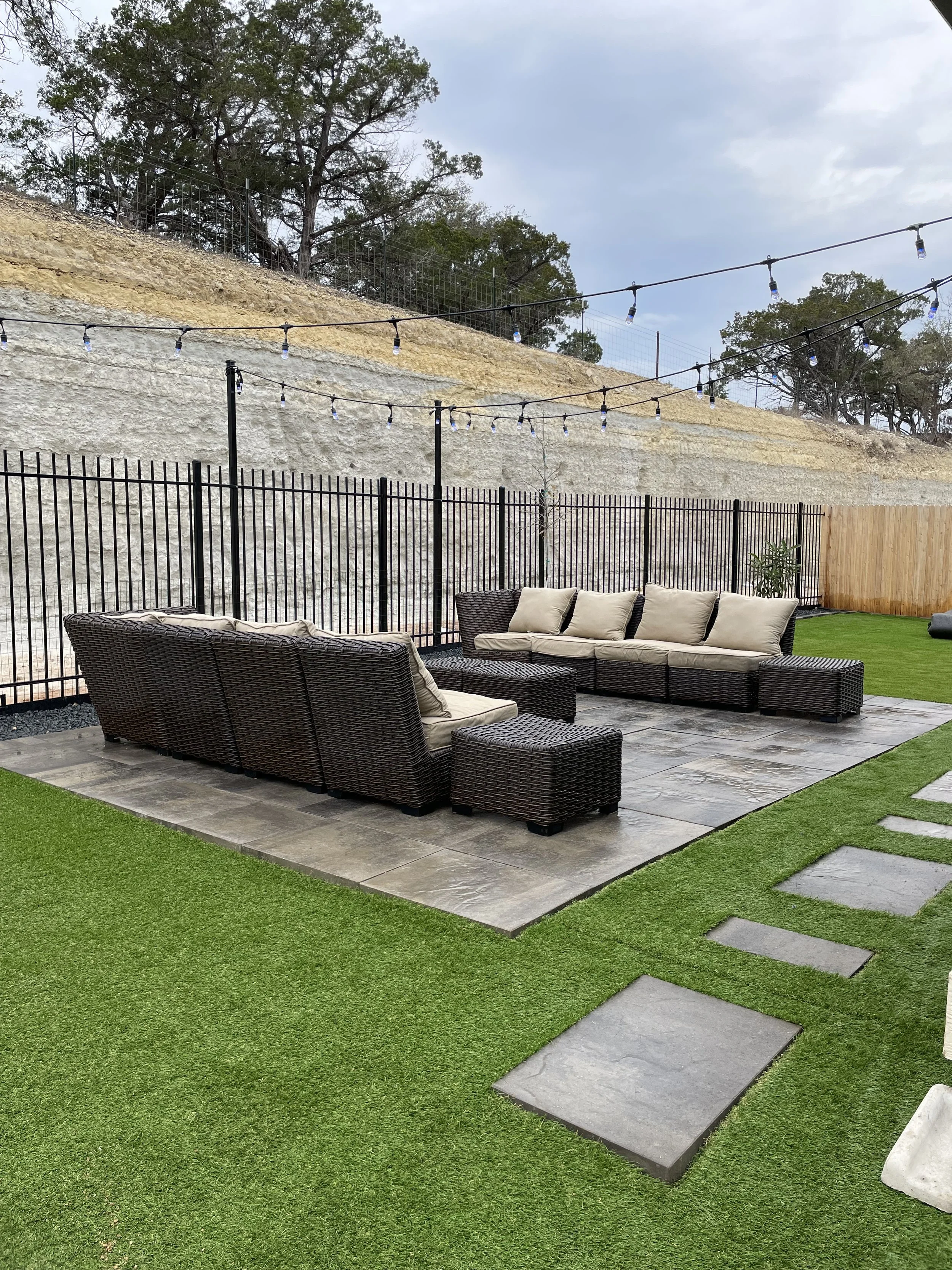 Outdoor patio area with wicker furniture, beige cushions, stone tile flooring, artificial grass, pathway with stepping stones, wooden fence, and string lights overhead, with a dirt hill and trees in the background.