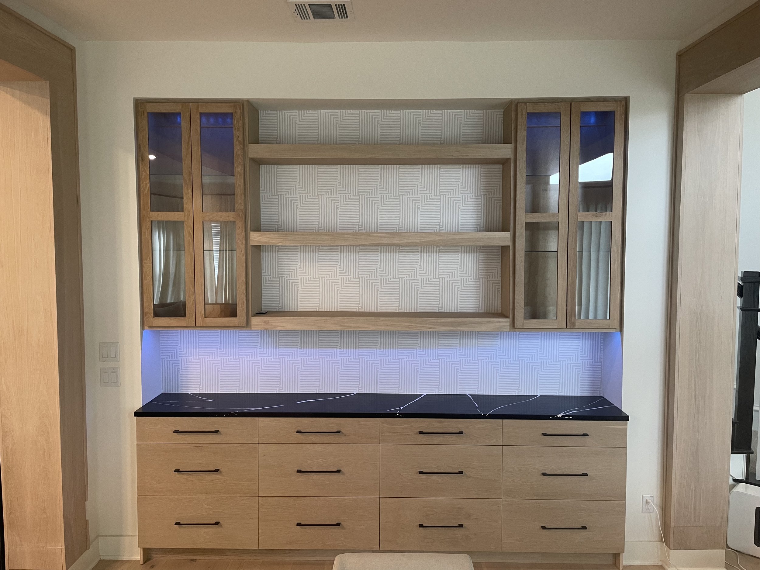 Image of a built-in wooden cabinet with glass doors on the top and drawers on the bottom, set against a wall with patterned wallpaper. The room has a ceiling vent and a power outlet visible.