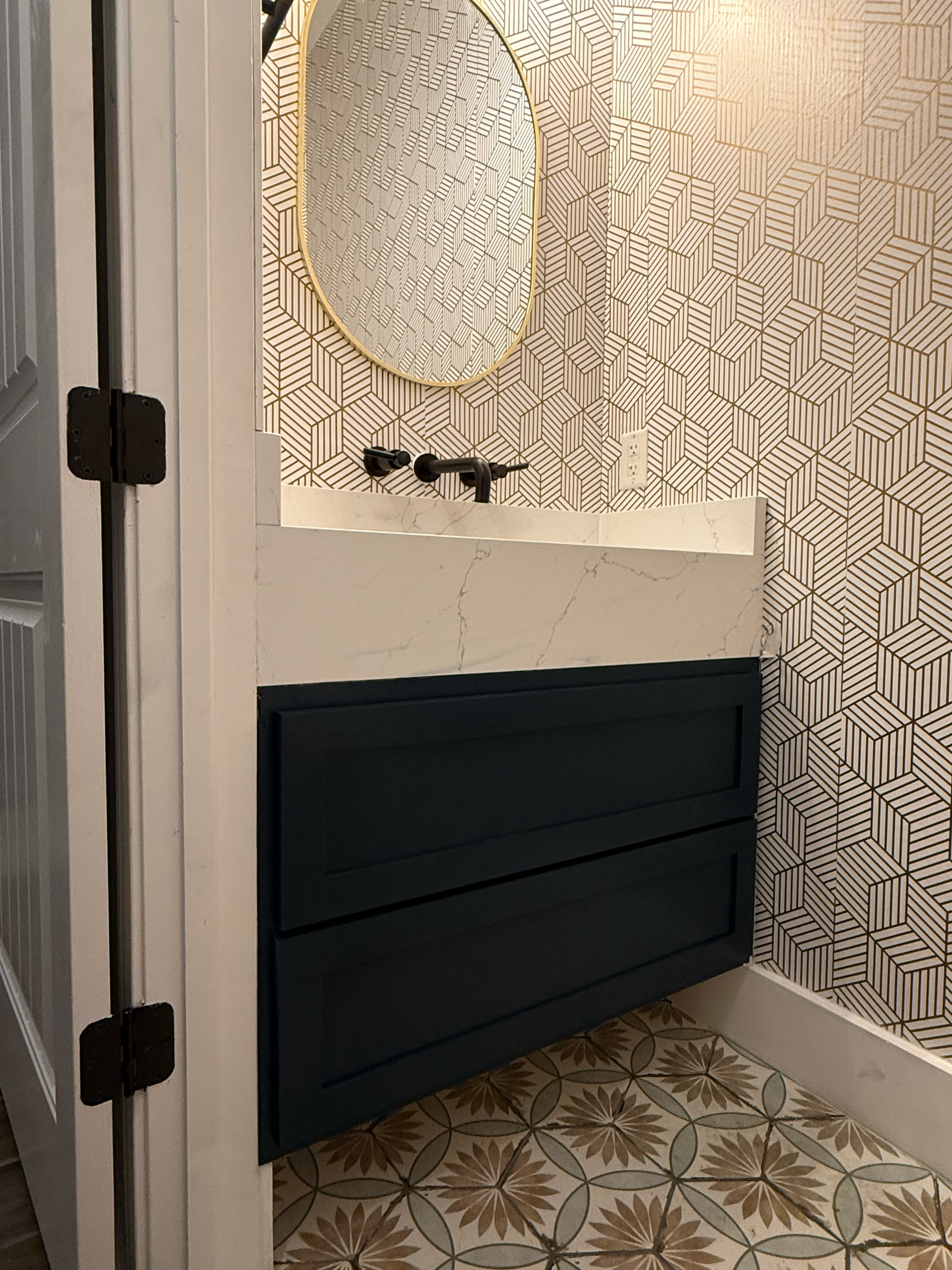 View of a bathroom vanity with a black cabinet, white marble countertop, black faucet, and a large mirror, with geometric patterned wallpaper and decorative floor tiles.