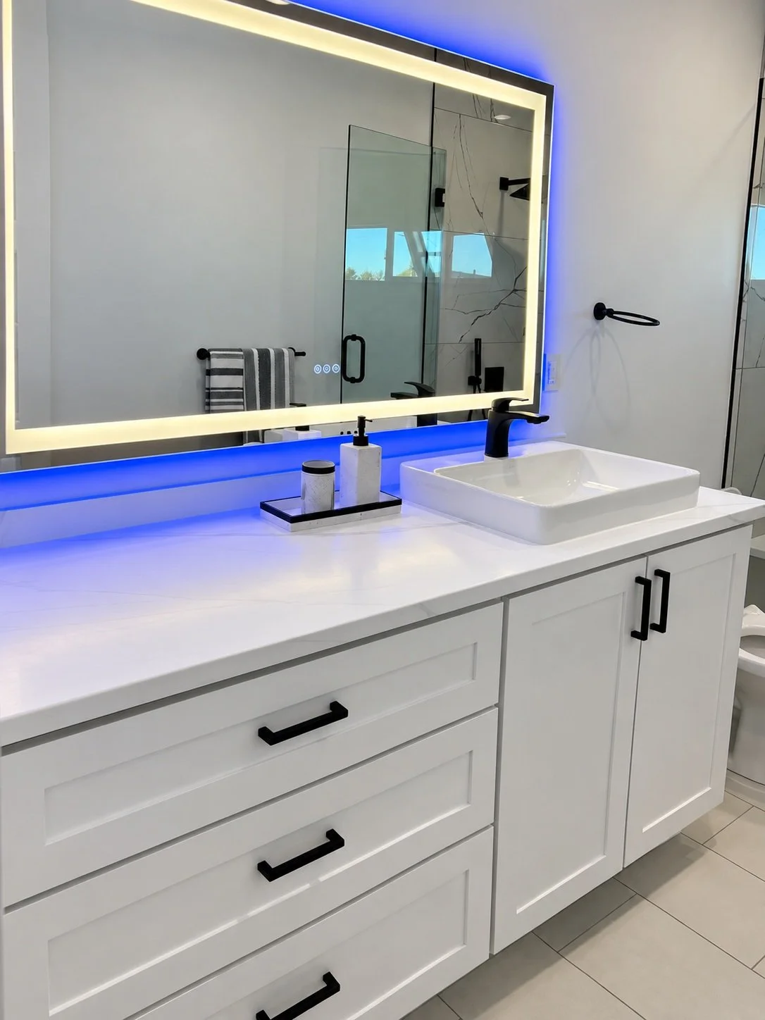 White vanity with black pulls, a rectangular vessel sink, black faucet, and a lighted mirror with blue accent lighting.