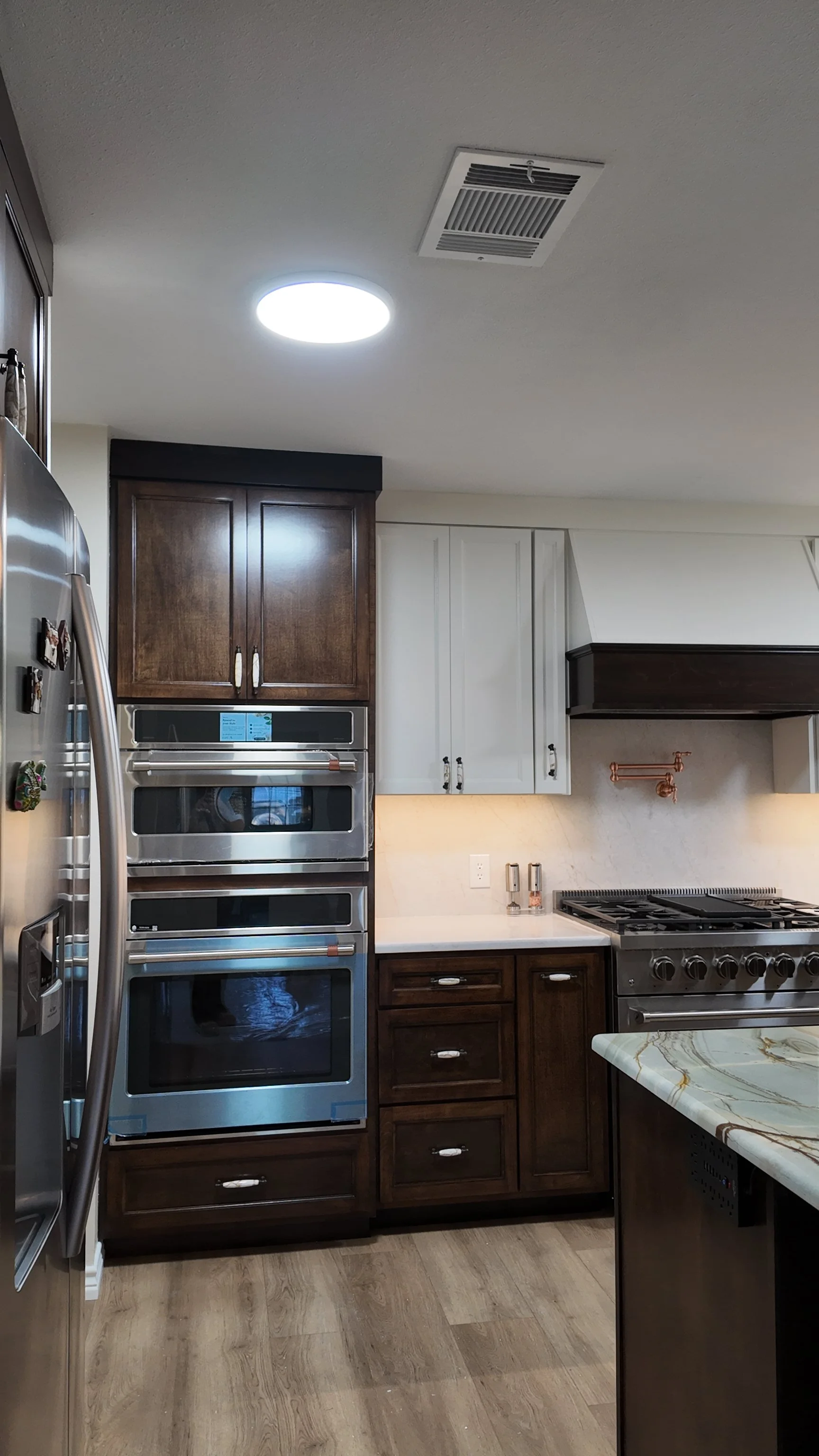 A modern kitchen with brown and white cabinets, a stainless steel oven, a gray stove, a marble countertop, and a ceiling light fixture.