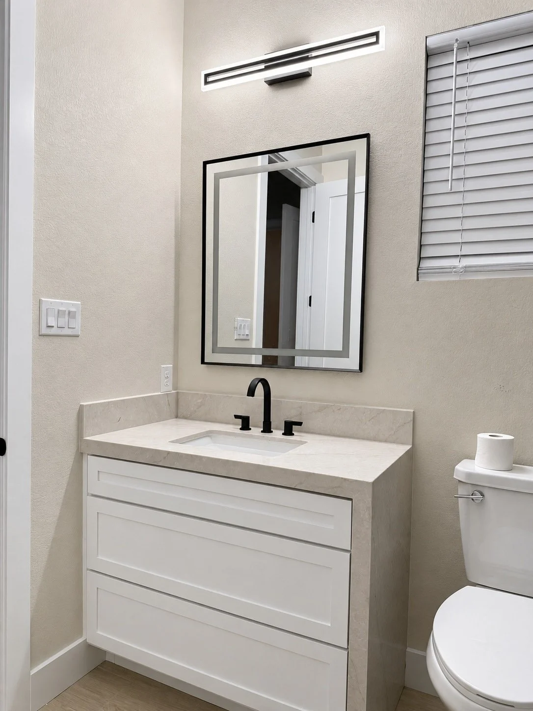 White single vanity with a light countertop, black faucet, framed mirror, and linear wall light.
