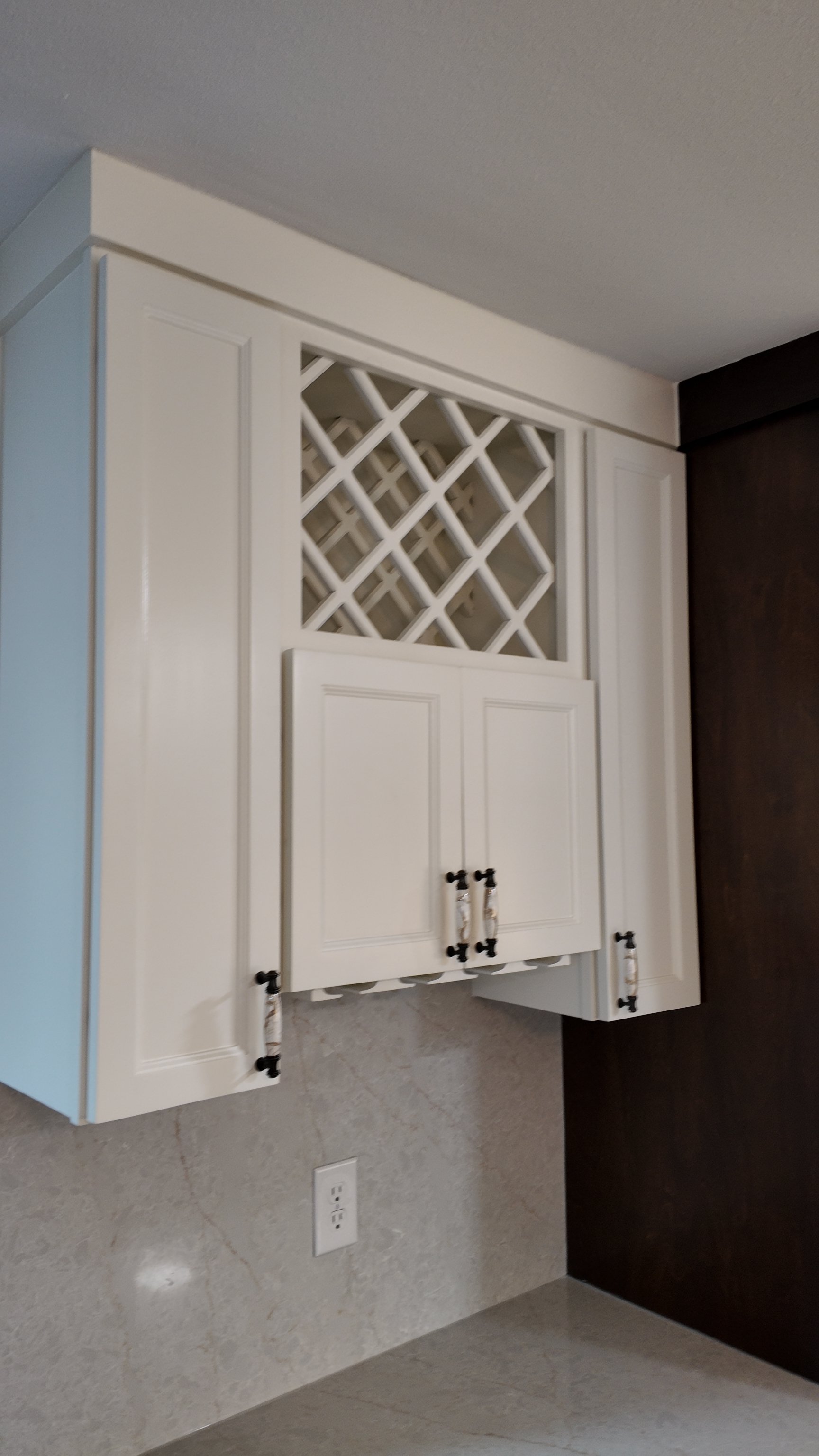 White kitchen cabinet with black handles, decorative glass wine rack, dark brown wall, electrical outlet on light-colored countertop, textured wall.