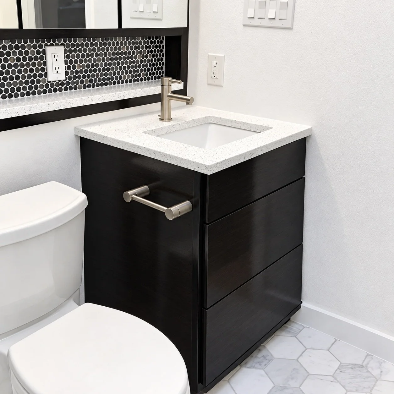 Compact dark vanity with a white countertop, brushed metal faucet, black penny-tile backsplash, and hexagonal floor tile.