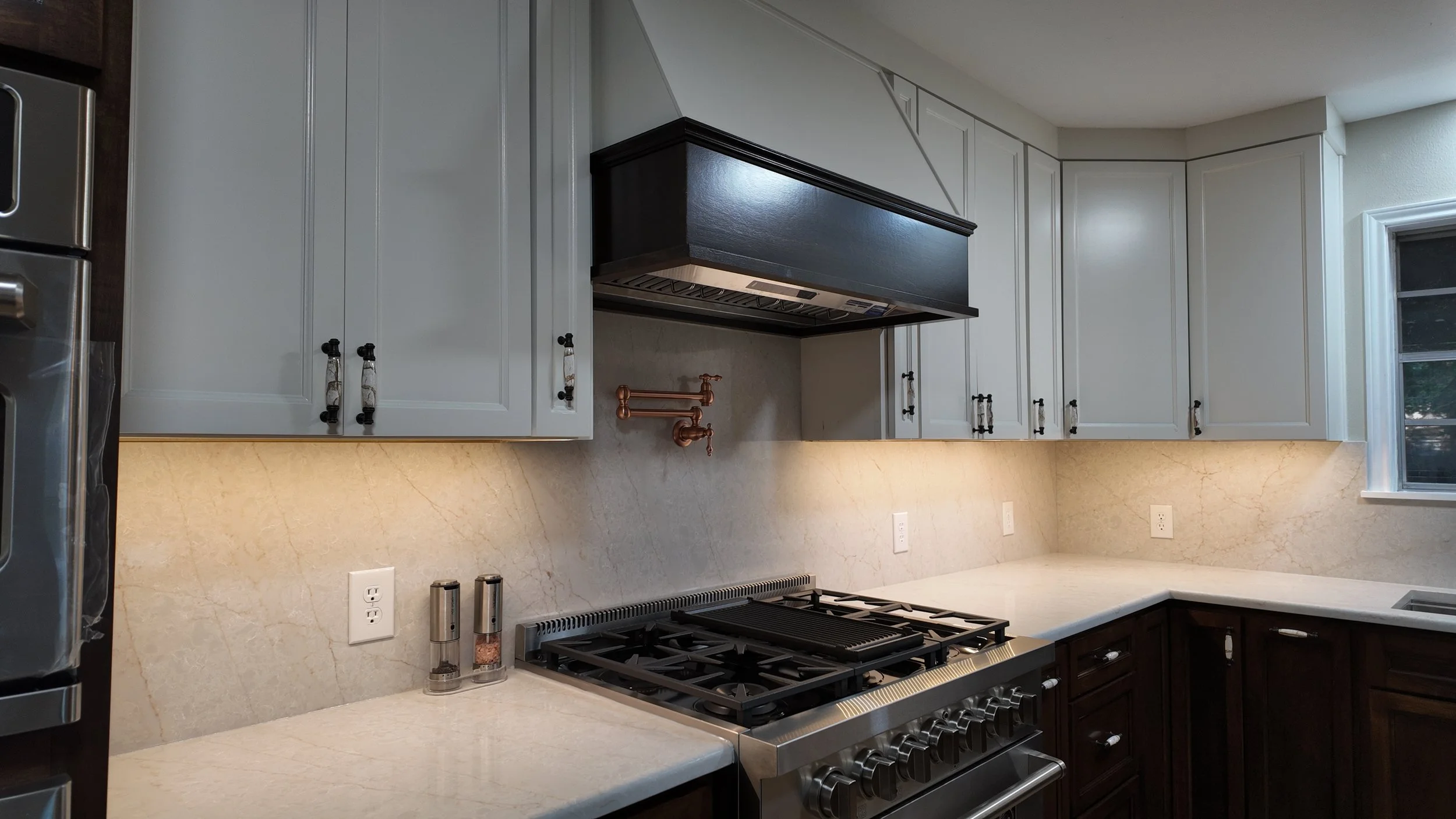 Kitchen with white cabinets, a black range hood above a stove, and a marble countertop. Copper pipes are installed on the wall. There are electrical outlets, salt and pepper shakers, and a window.