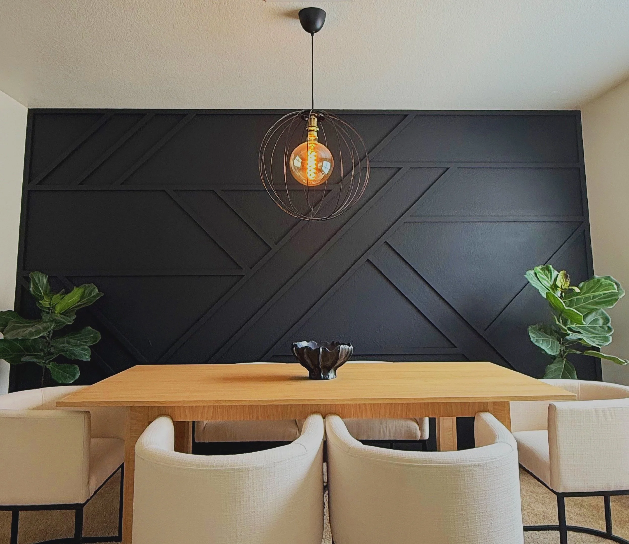 A wooden dining table with a black bowl in the center, surrounded by white cushioned chairs. Green plants are positioned on either side of the table. Behind the table is a black, geometric-patterned wall, and a modern pendant light with an orange, spherical filament bulb hangs above the center of the table.