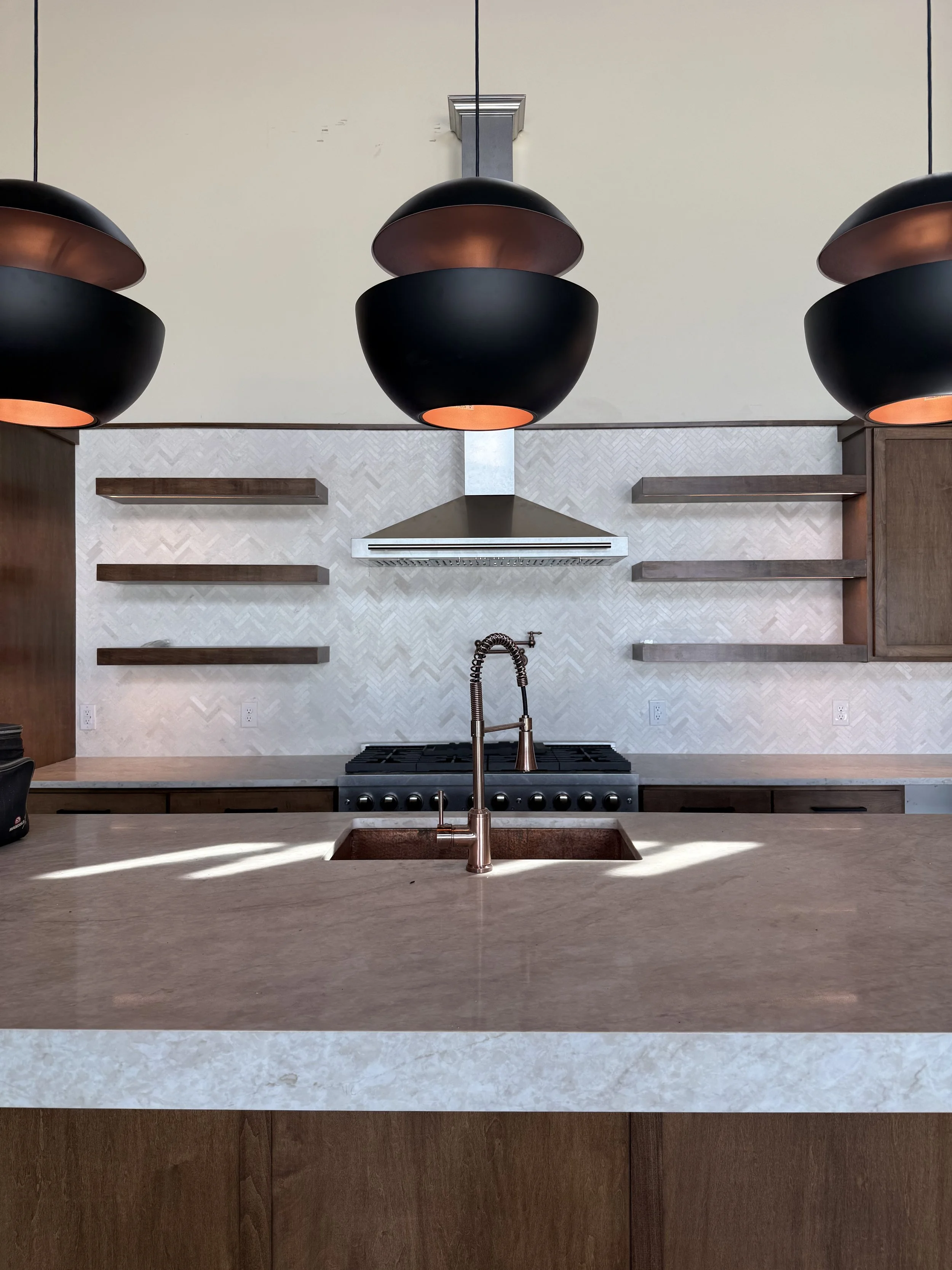 Modern kitchen with beige countertop, copper-colored sink faucet, beige backsplash, and dark pendant lights hanging from a white ceiling; wooden shelving on the wall behind, and a stove beneath a metallic range hood.