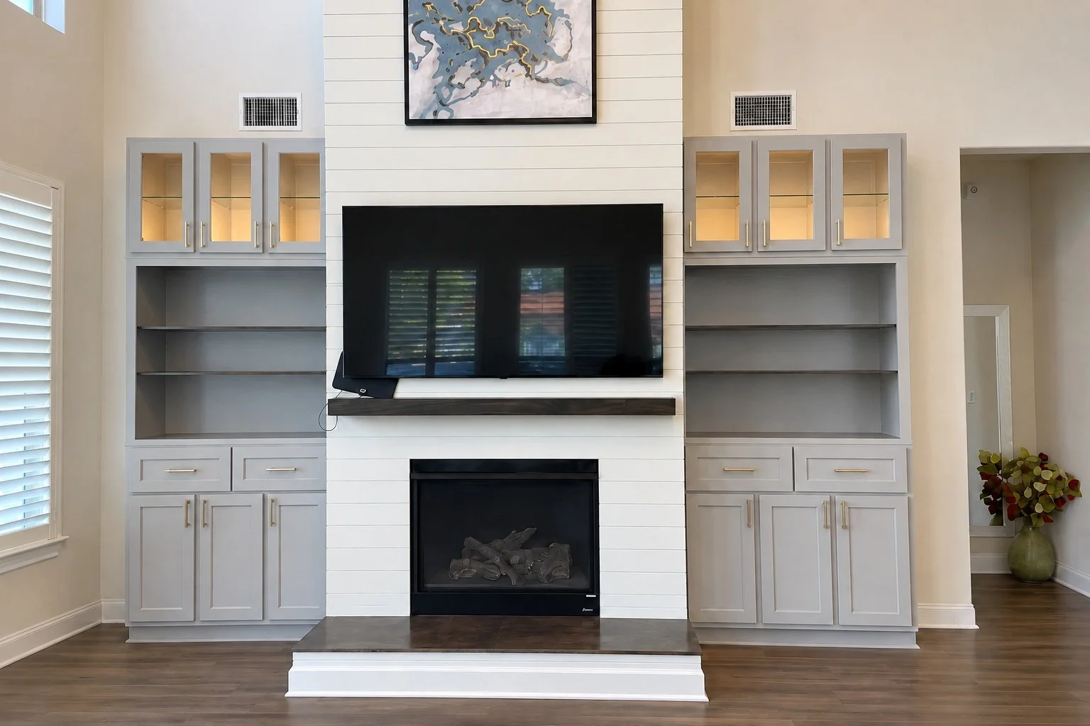 Gray display cabinets and open shelving frame a white shiplap fireplace wall, with glass-front doors, warm lighting, and gold-tone pulls.