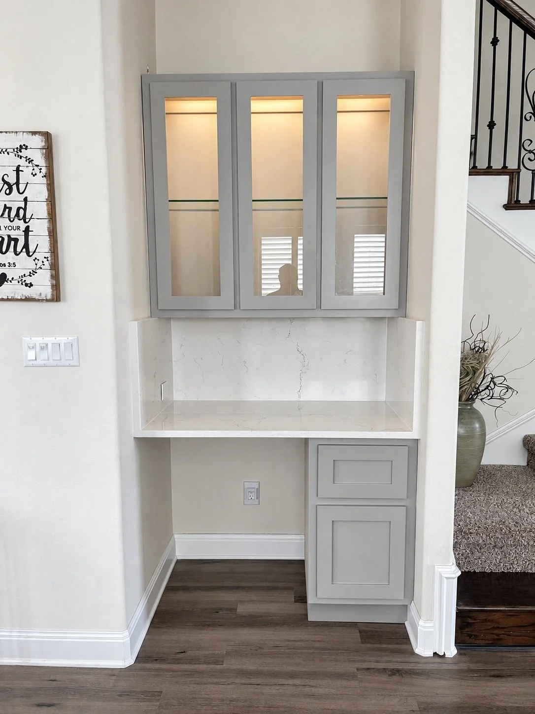 Compact gray built-in nook with illuminated glass-front upper cabinets, a pale work surface, and lower storage.