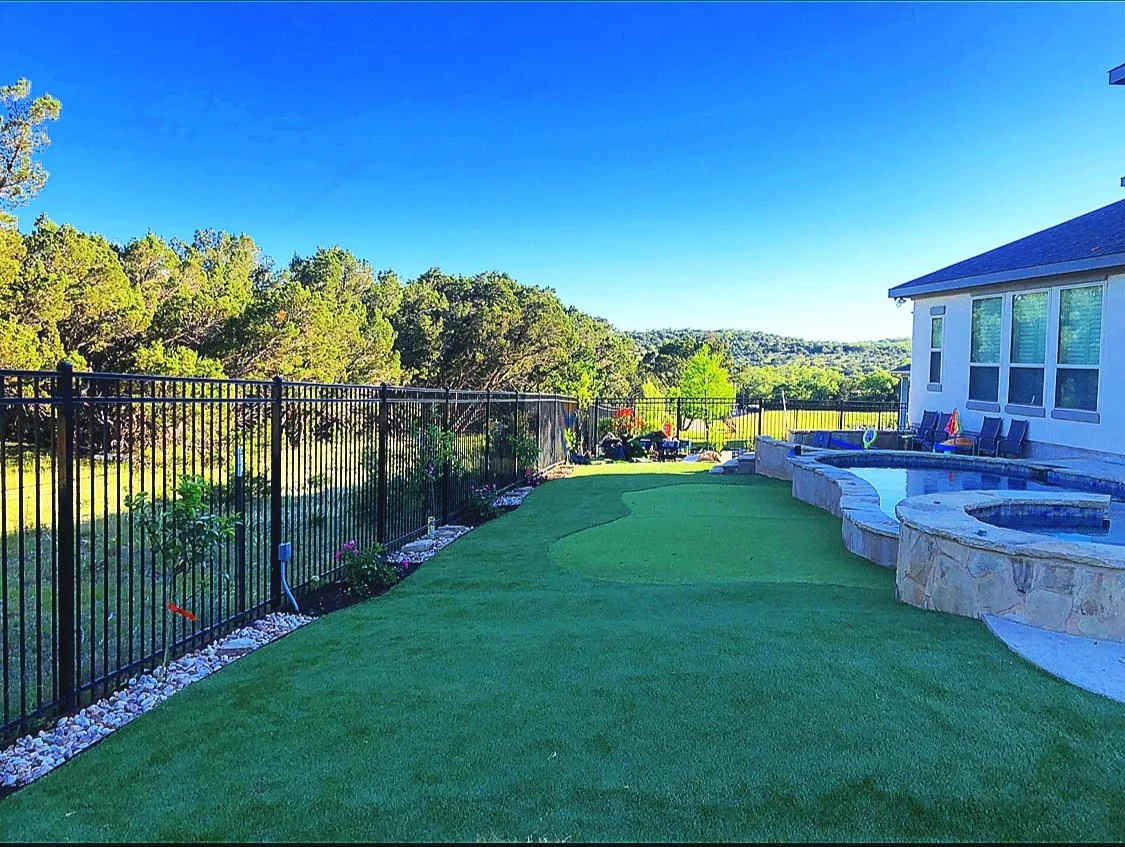 Backyard with artificial grass, a pool, outdoor furniture, and a black metal fence, with trees and a clear blue sky in the background.