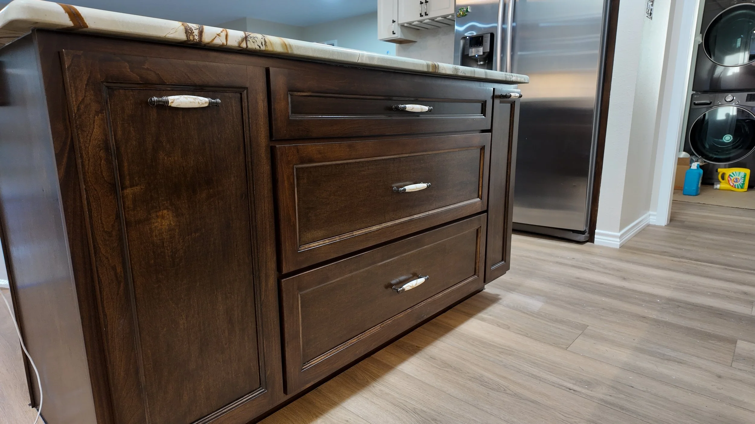 Dark wooden kitchen island with a light marble countertop in a modern kitchen, with stainless steel refrigerator and laundry appliances in the background, and cleaning supplies on the floor.
