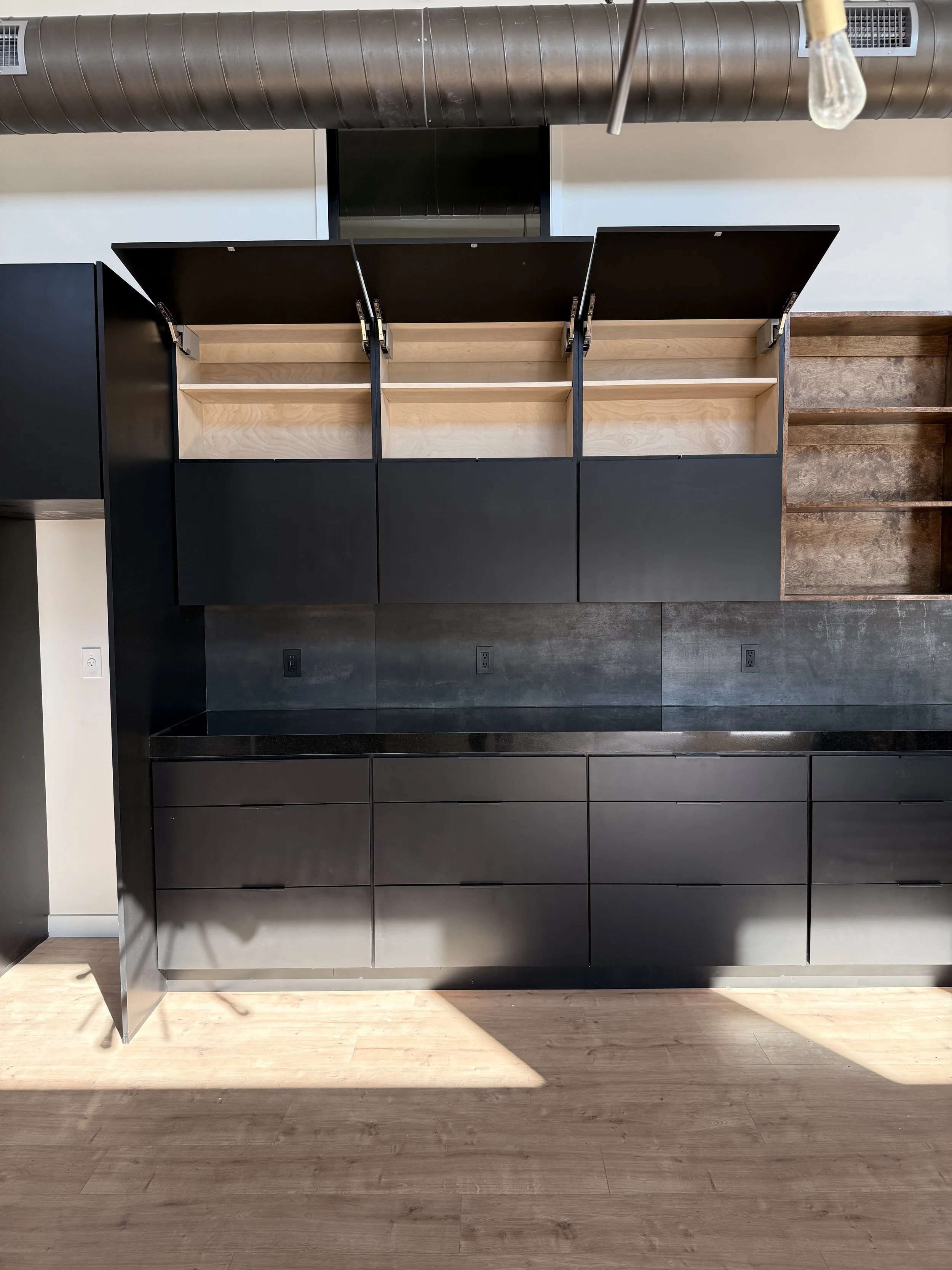 An unfinished modern kitchen cabinet with black exterior and open upper shelves, mounted on a wall in a room with wooden flooring and an exposed duct ceiling. A light bulb hangs from the ceiling.
