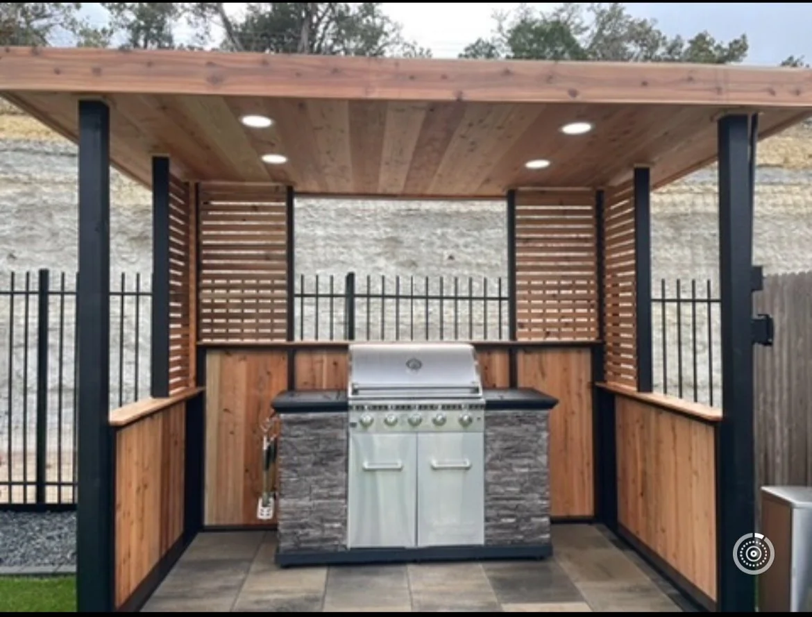 Outdoor kitchen with a built-in barbecue grill, wooden and black metal fencing, and a slatted wooden roof with recessed lighting.