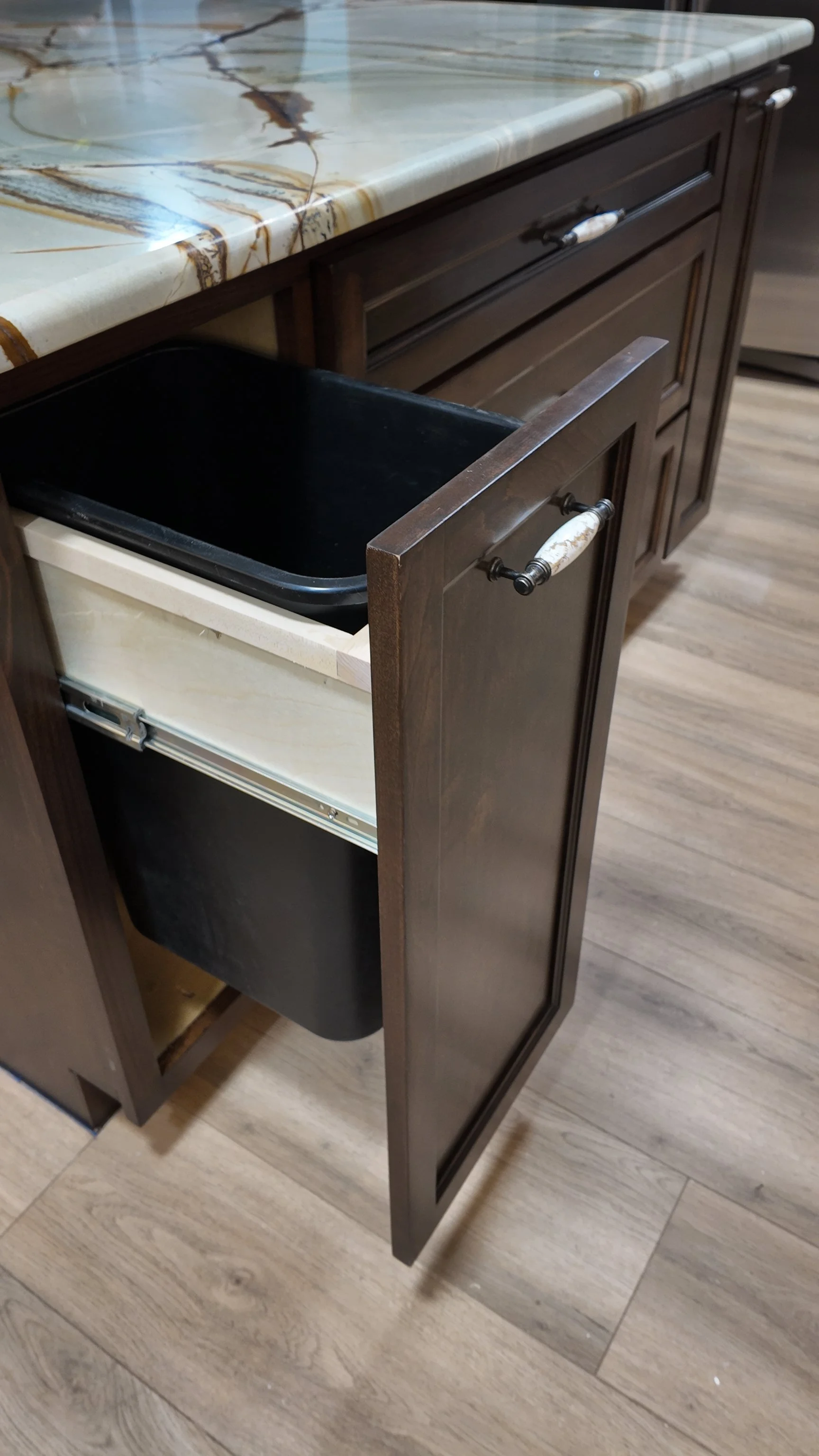 Open kitchen cabinet with a marble countertop, revealing an empty black trash bin inside the lower compartment.