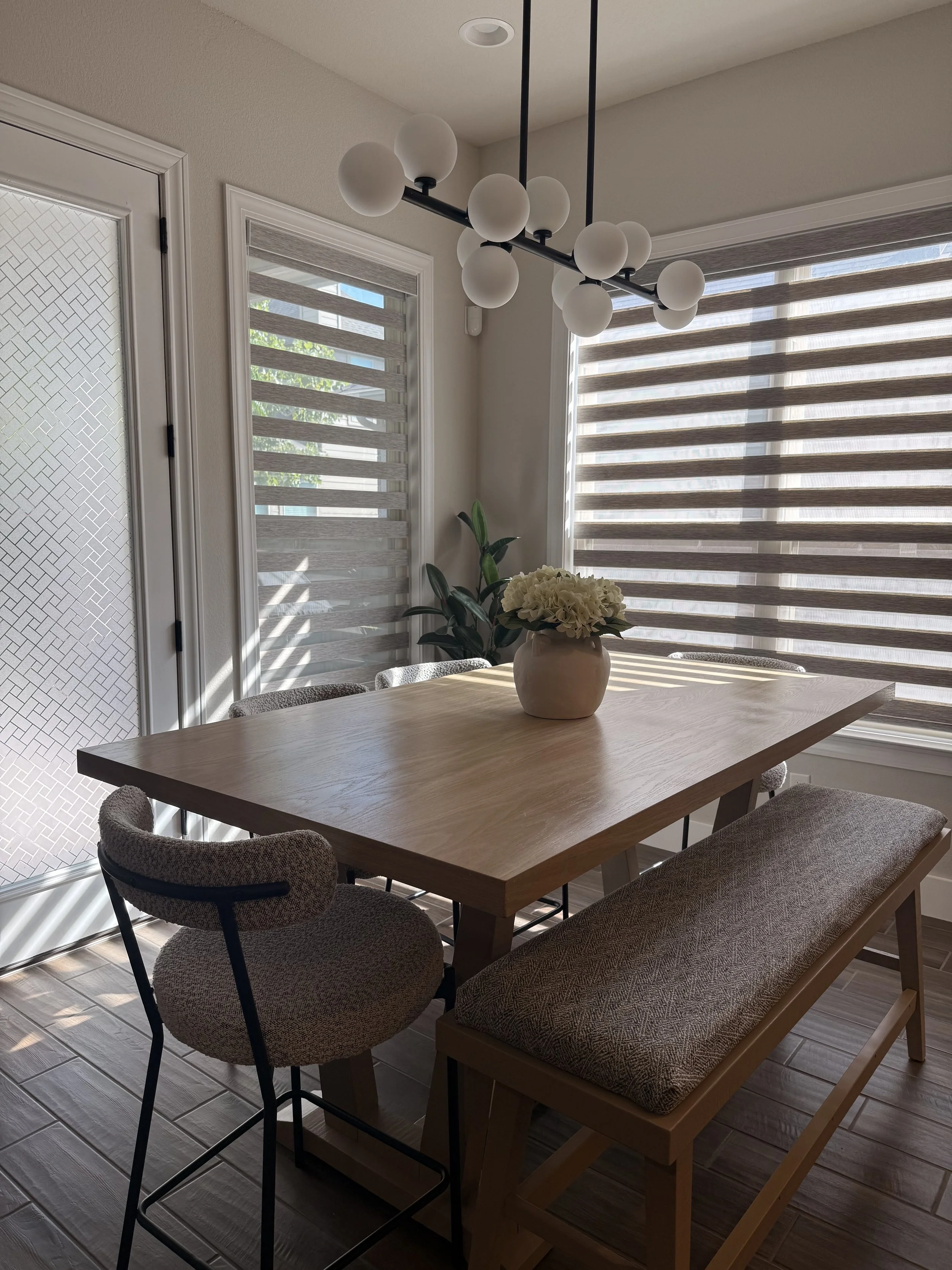 A dining room with a wooden table, a ceramic vase with white flowers, and six chairs. Large windows with blinds allow sunlight to brighten the room.