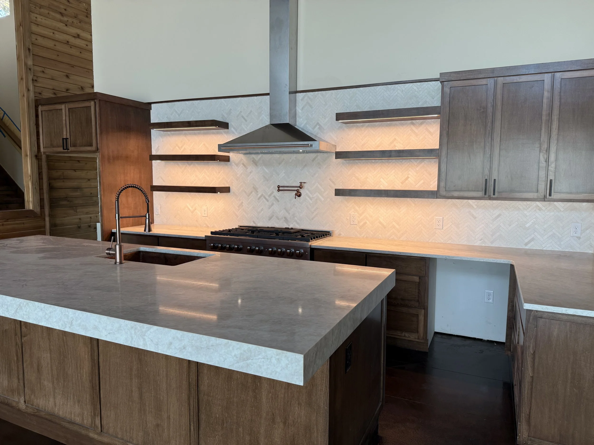 Modern kitchen with wooden cabinets, a stainless steel range hood, a stove, a black faucet, and a marble countertop. Open shelving and tiled backsplash are also visible.