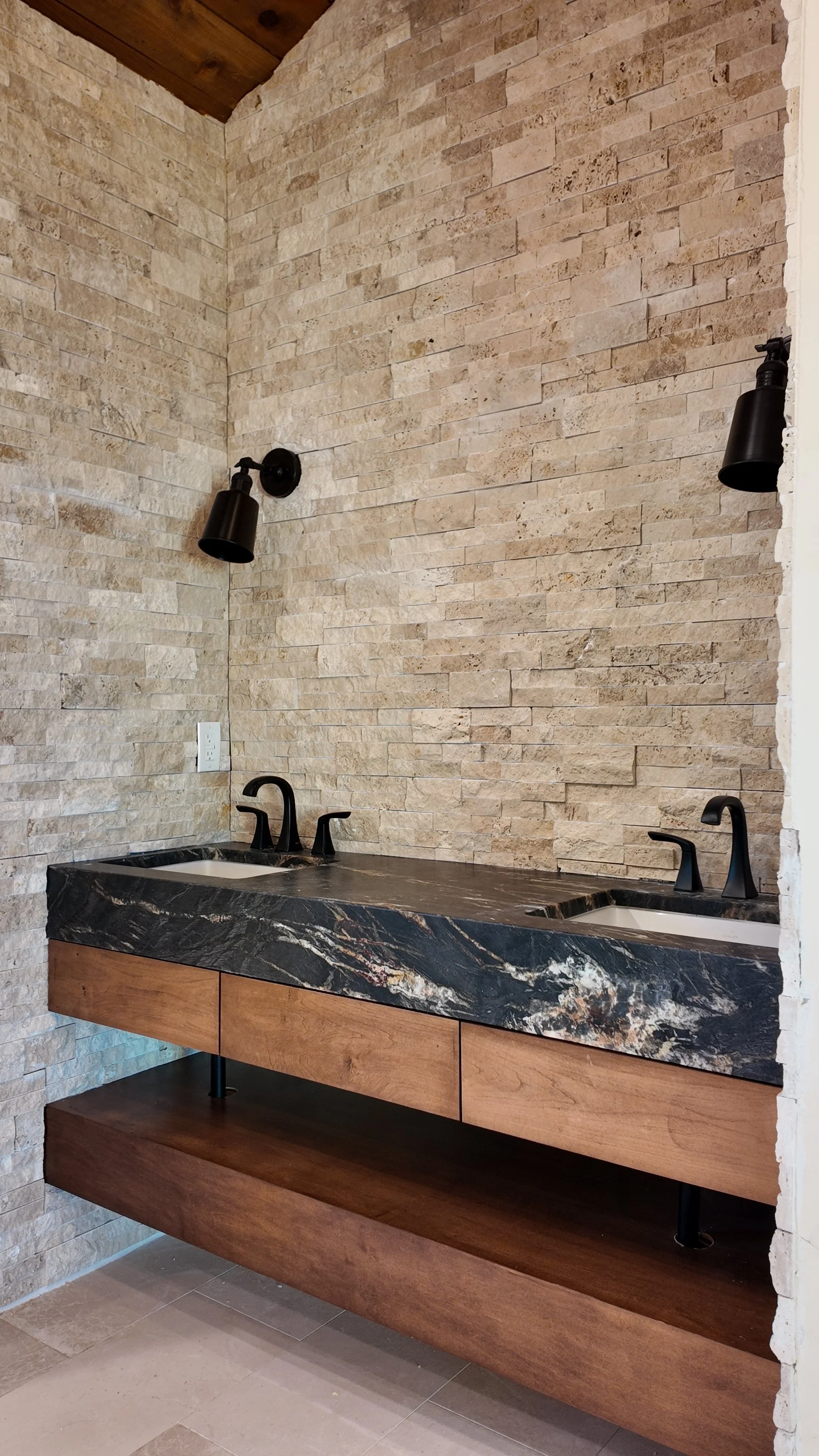 Double vanity with black faucets and a black marble countertop, set against a beige brick wall with two black wall-mounted light fixtures.