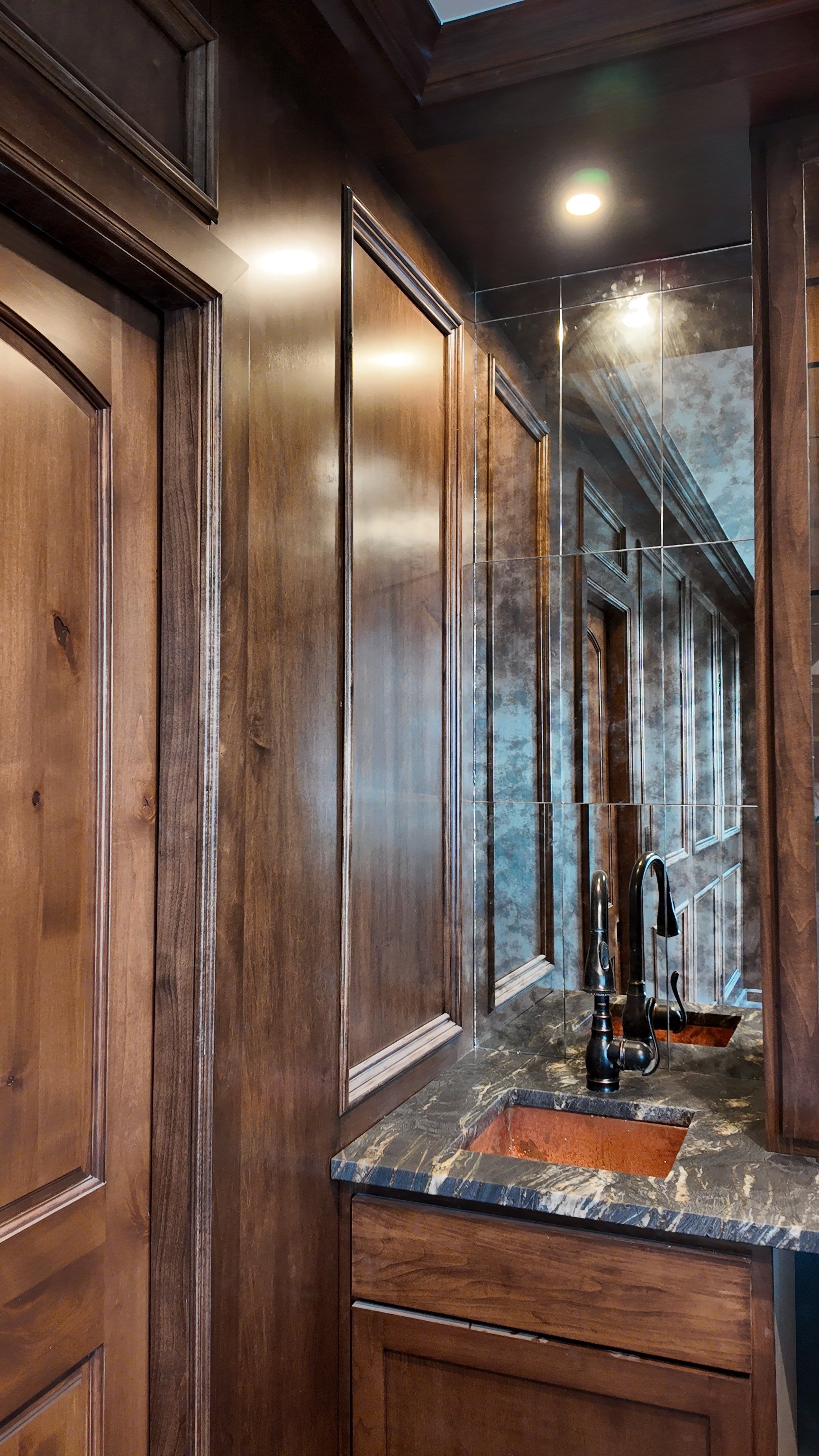 A kitchen or bathroom area with wooden cabinetry, a double sink with a dark bronze faucet, and a large mirror reflecting the cabinets and ceiling.