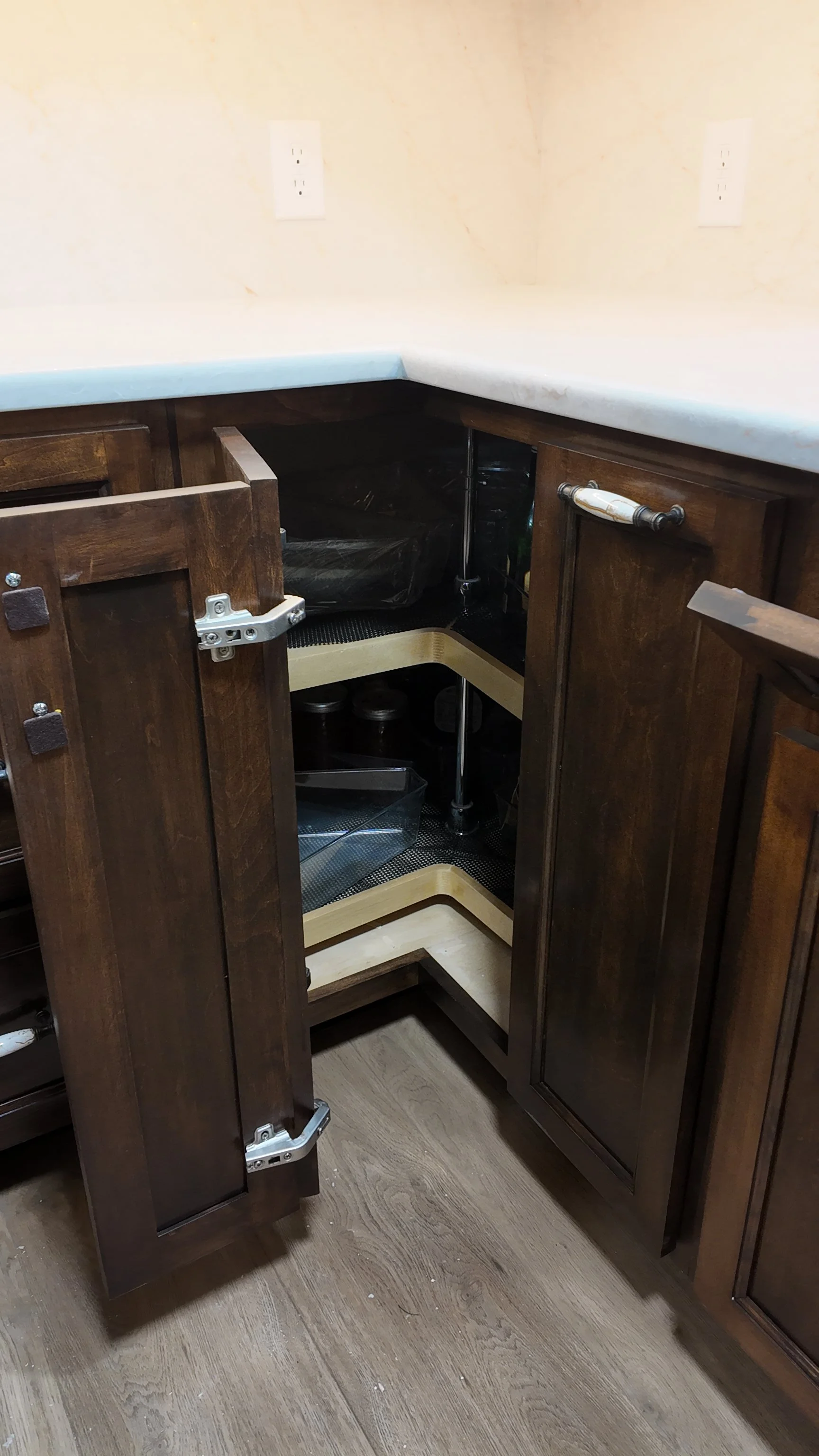 Unfinished dark wood kitchen cabinet with open doors, empty shelves, and visible hardware under a white countertop with two electrical outlets on the wall above.