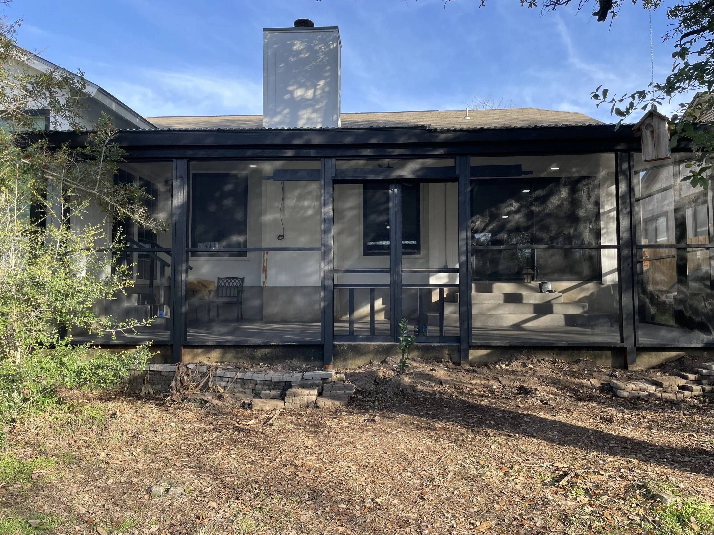 Backyard view of a house with a screened porch and a chimney, trees and blue sky in the background, bare soil and scattered bricks in the foreground.