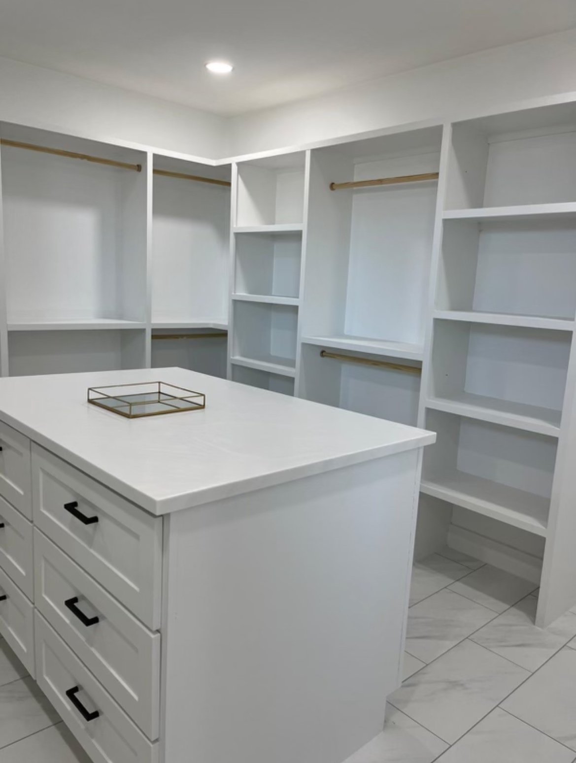 Empty white walk-in closet with built-in shelving and hanging rods, central island with drawers, and a tray on top.