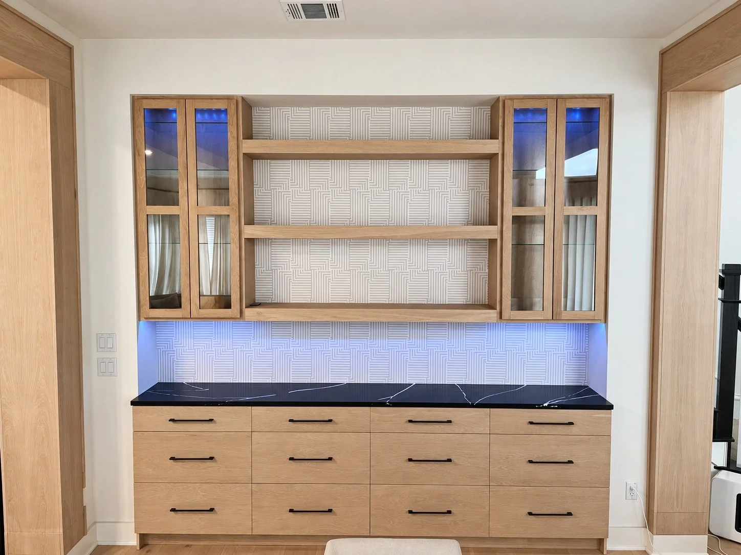 Natural wood bar cabinetry with glass upper doors, a dark veined countertop, black pulls, and blue accent lighting.
