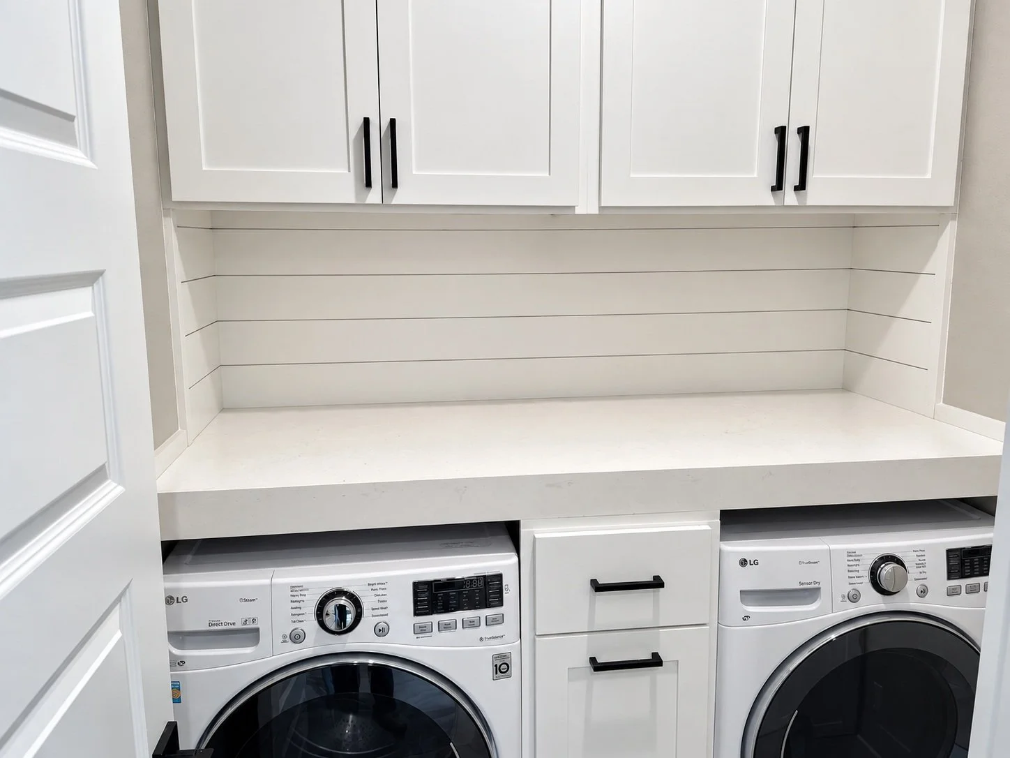 White laundry-room built-ins with black cabinet pulls, a continuous countertop, and drawers between the washer and dryer.
