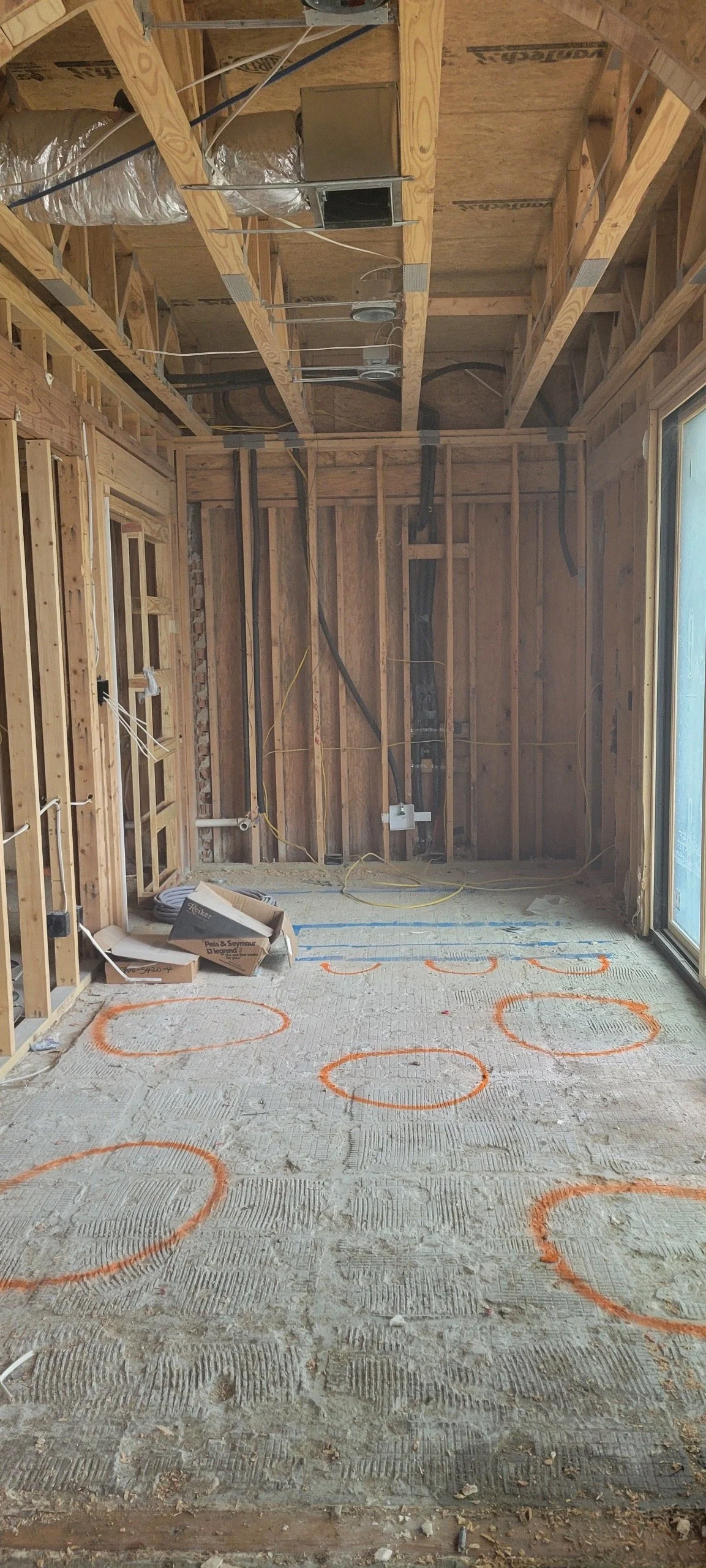 Interior of a room under construction with exposed wooden framing, electrical wiring, and orange circles marked on the concrete floor.