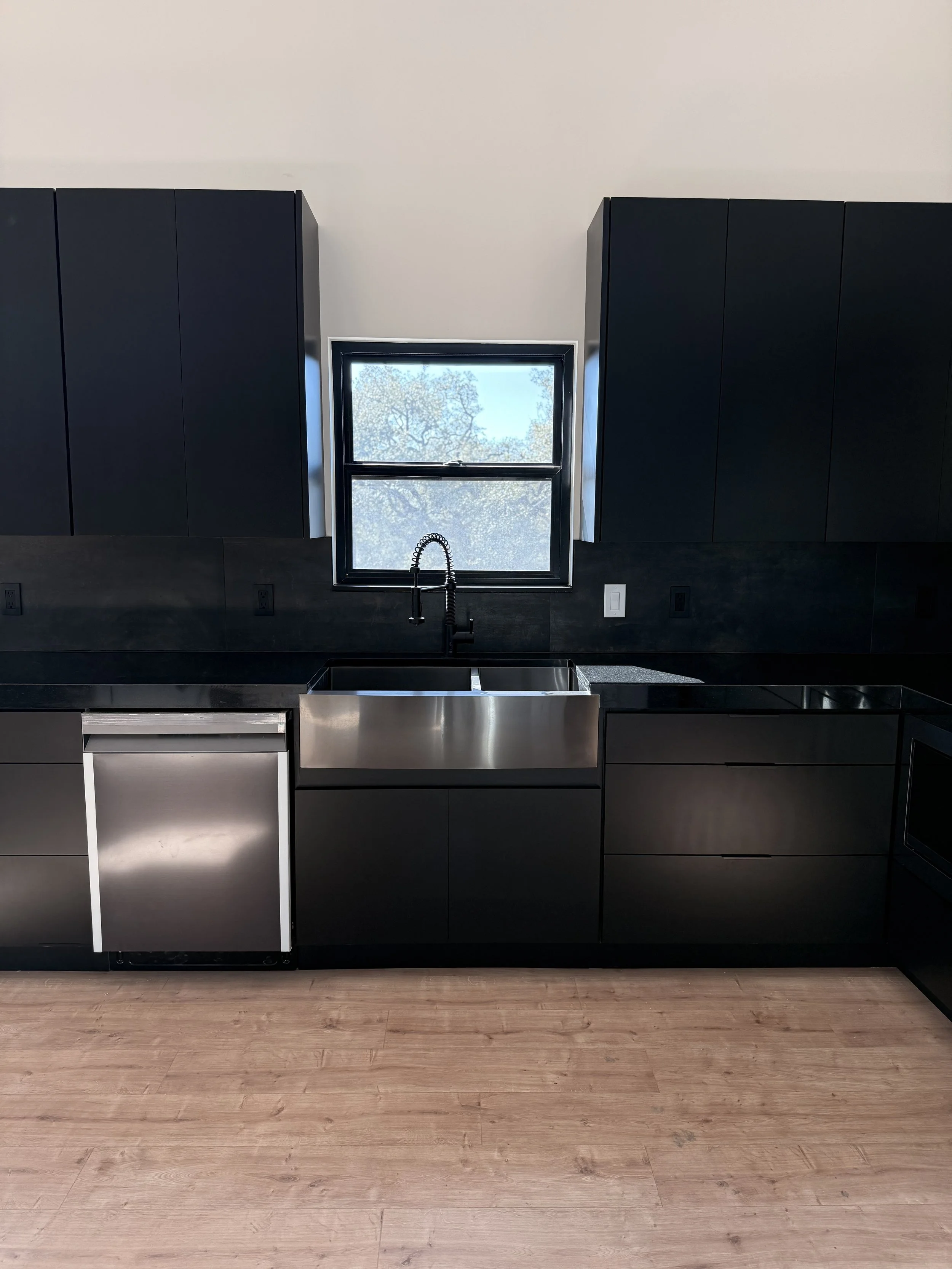 Modern kitchen with black cabinets, a stainless steel dishwasher, a window above the sink, and a wooden floor.