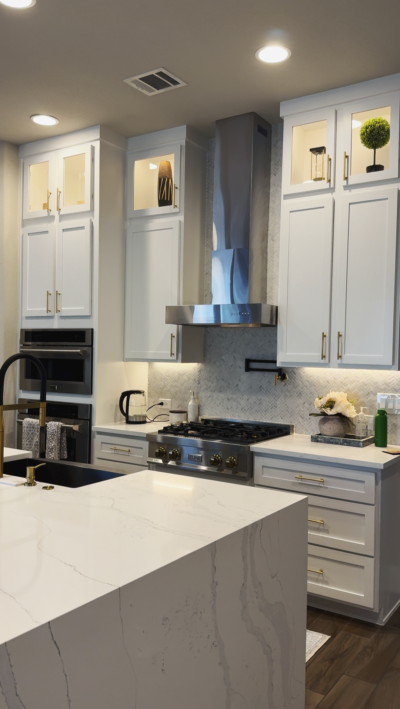 Modern kitchen with white cabinets, stainless steel range hood, marble countertop, and decorative items such as potted plants and flowers.