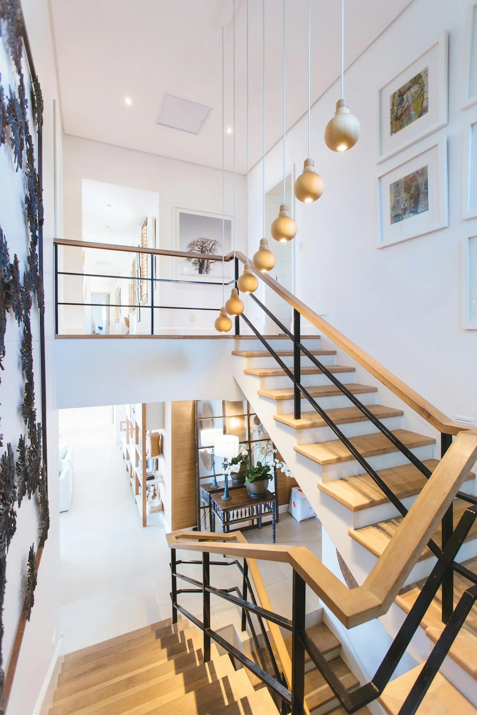 Modern interior staircase with wooden steps and black metal railings, hanging pendant lights, wall art, and framed pictures on a light-colored wall.