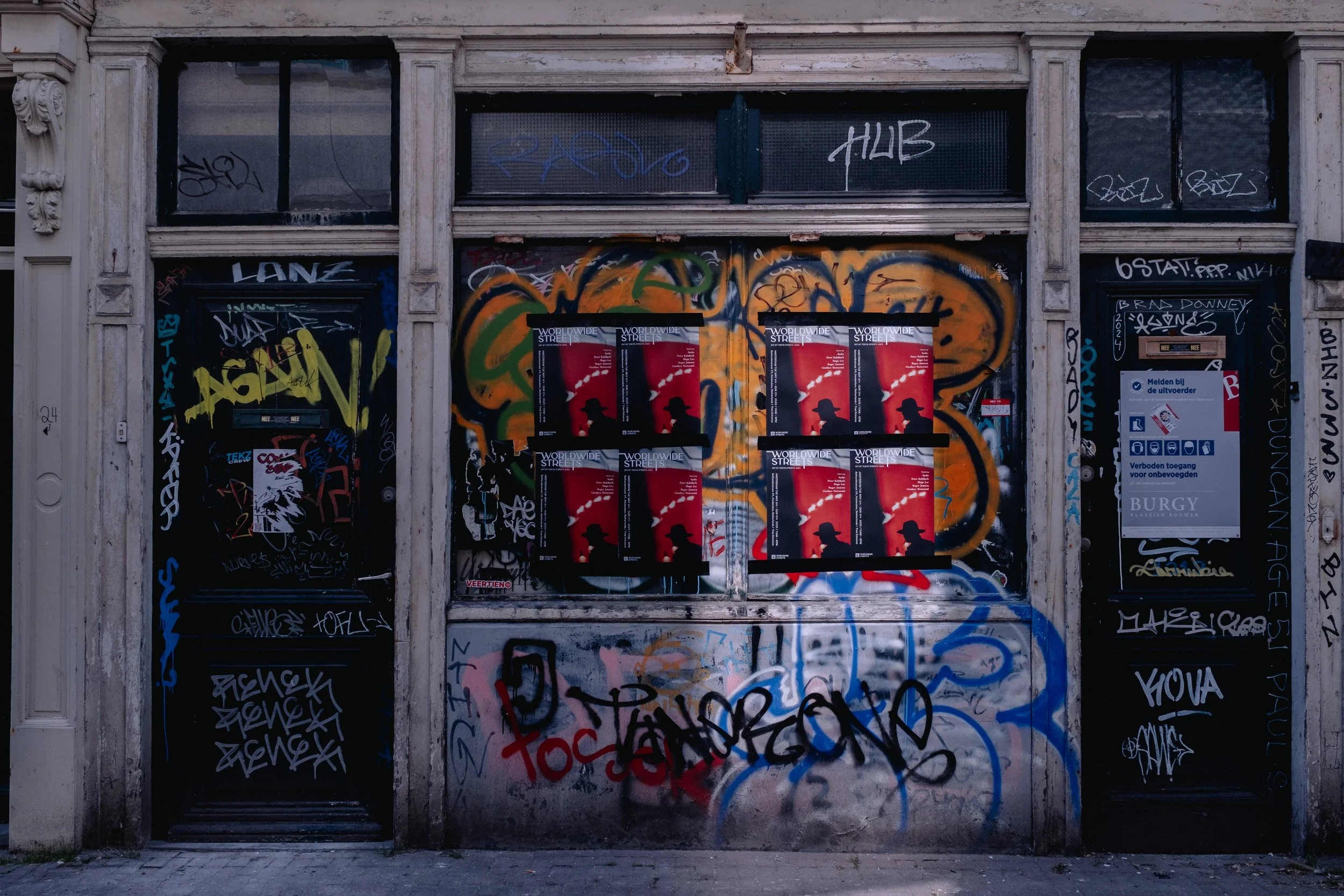 A storefront covered in colorful graffiti with multiple black posters showing a silhouette of a man wearing a hat and smoke. The posters read "Worldwide Streets." The graffiti includes tags, scribbles, and a large orange and green spray-painted desig
