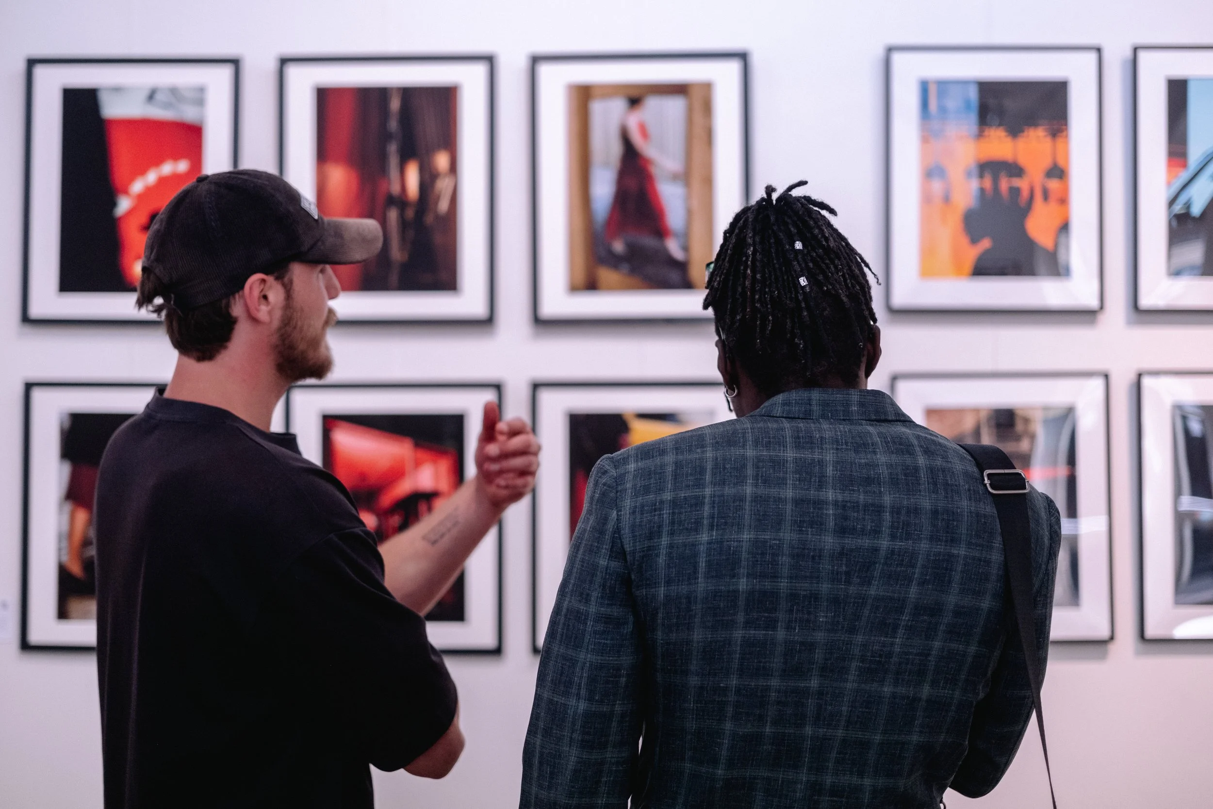 Two men viewing artwork in an art gallery, with several framed photographs on the wall behind them.