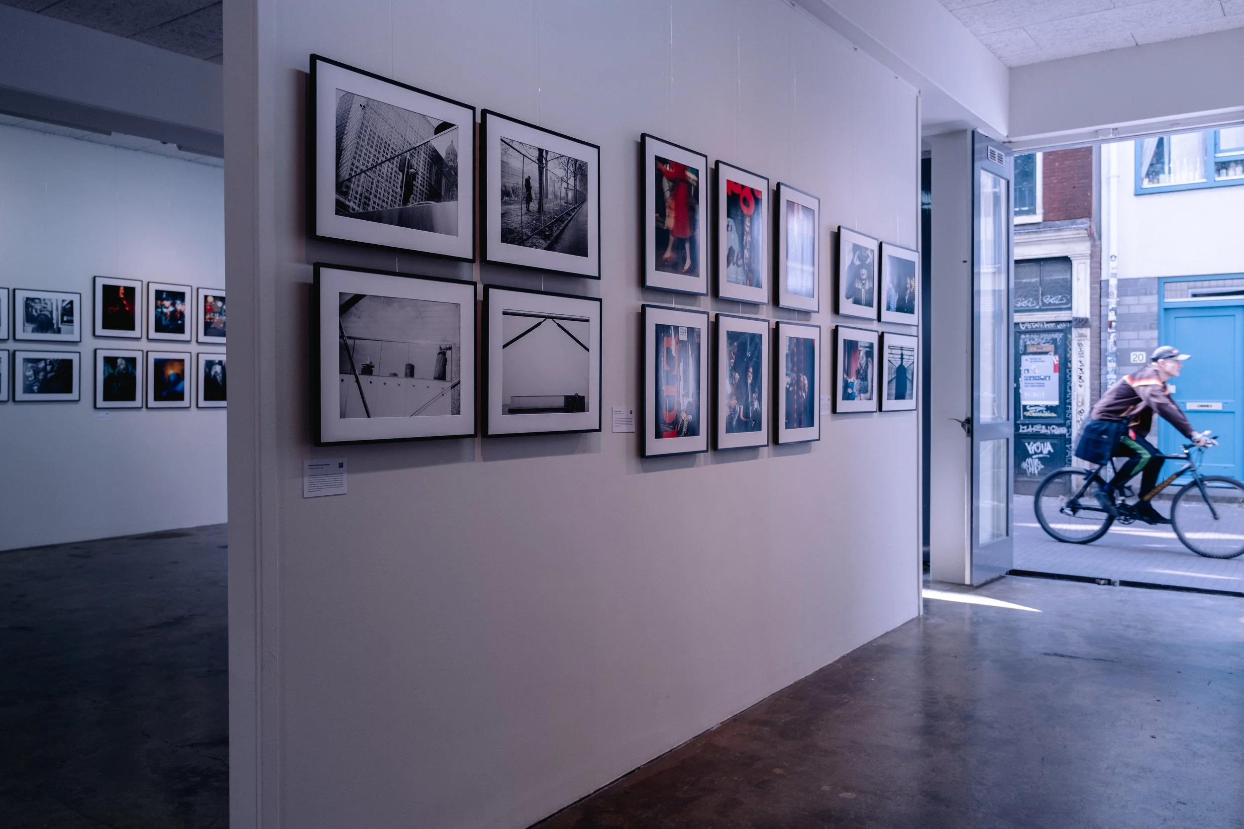 Street Photography art gallery in Amsterdam with framed photographs displayed on white walls, featuring silhouettes, reflections, and urban scenes, with a partly visible art desk and chairs in background.
