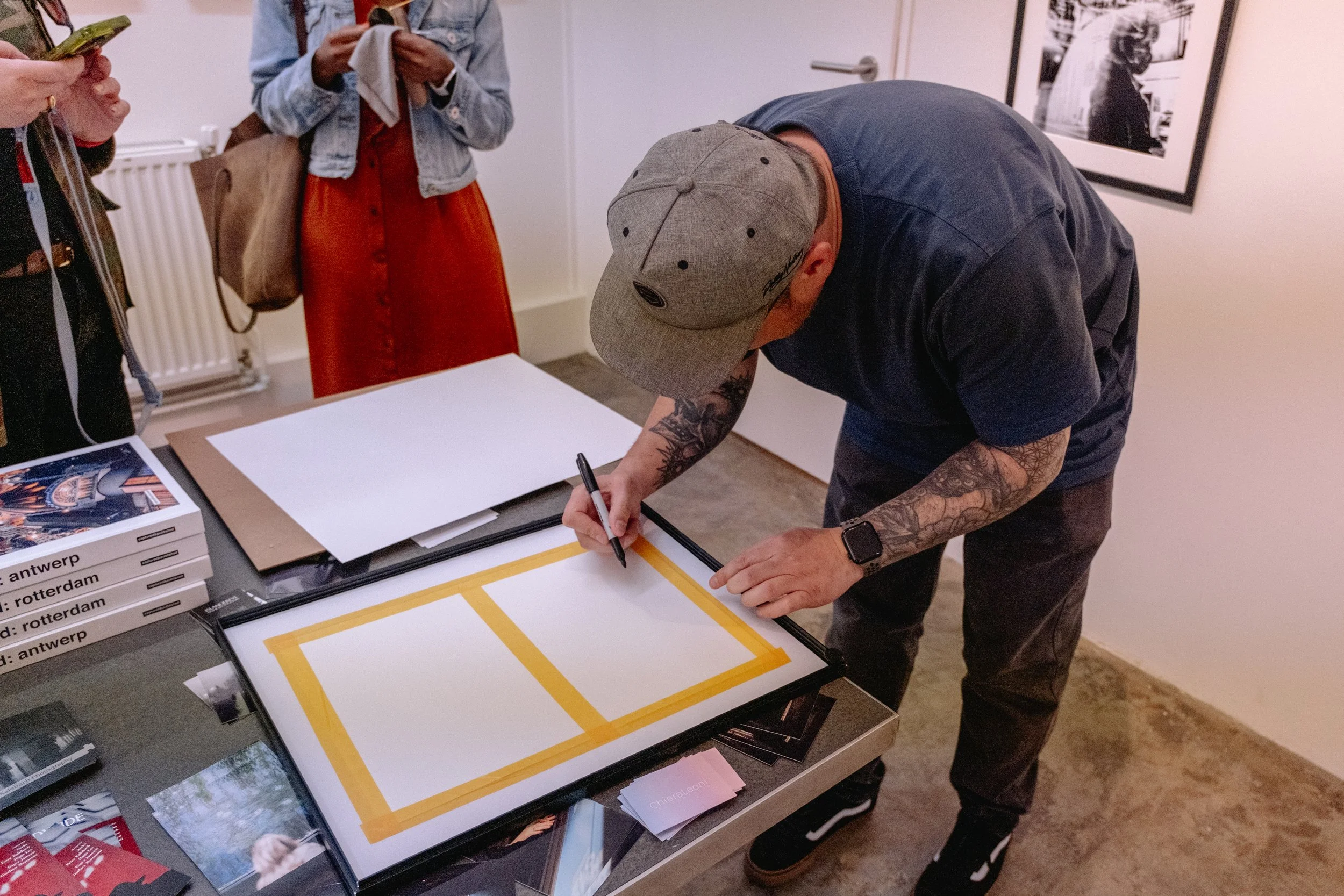 Man with tattoos signing a picture frame at an event table, with two women standing nearby, one using a phone and the other holding a folding paper, in an art gallery setting.