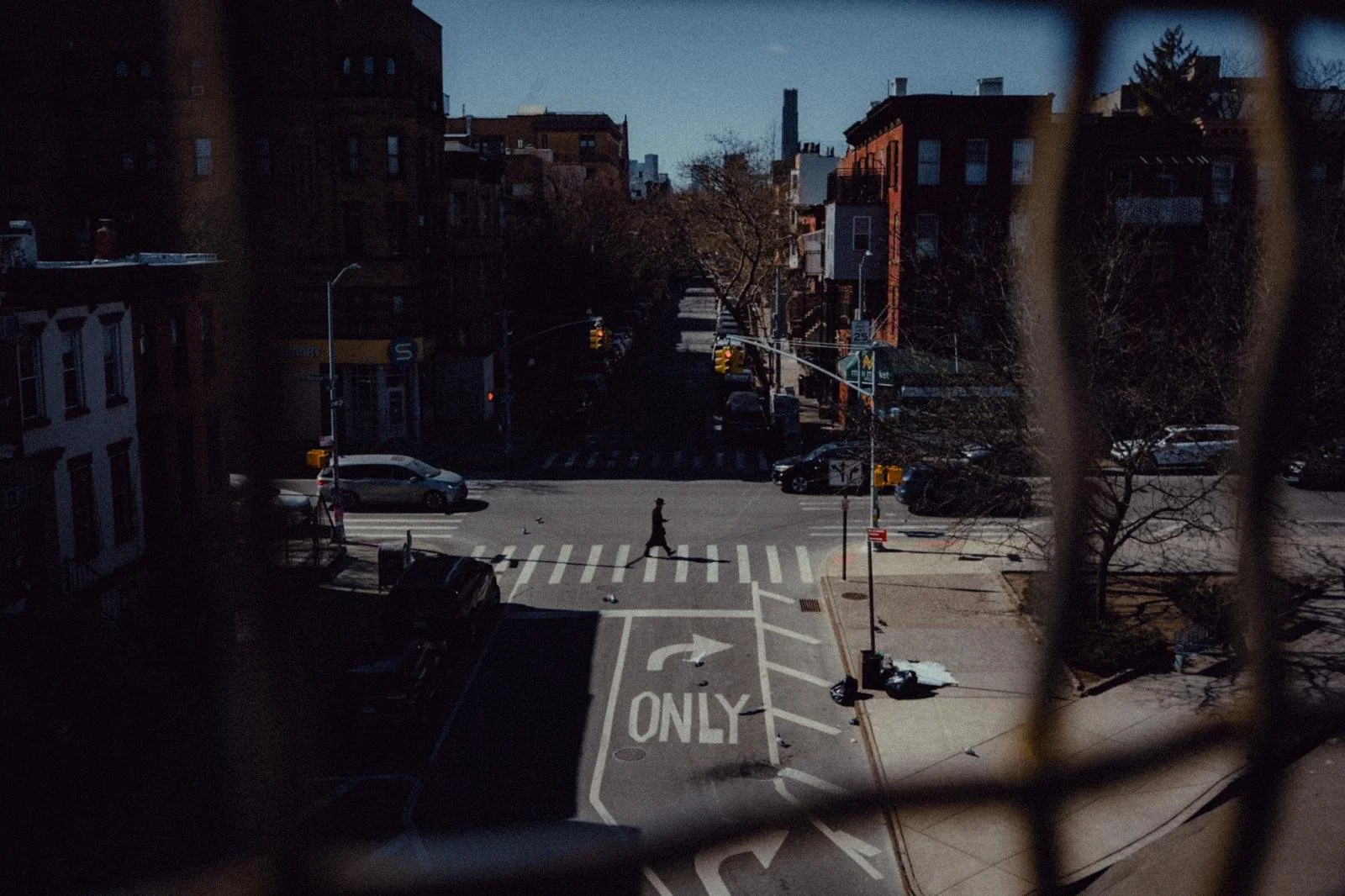 A city street viewed from a balcony or window, with a person crossing at a crosswalk, cars on the road, and buildings in the background, framed by a safety railing or bars in the foreground.