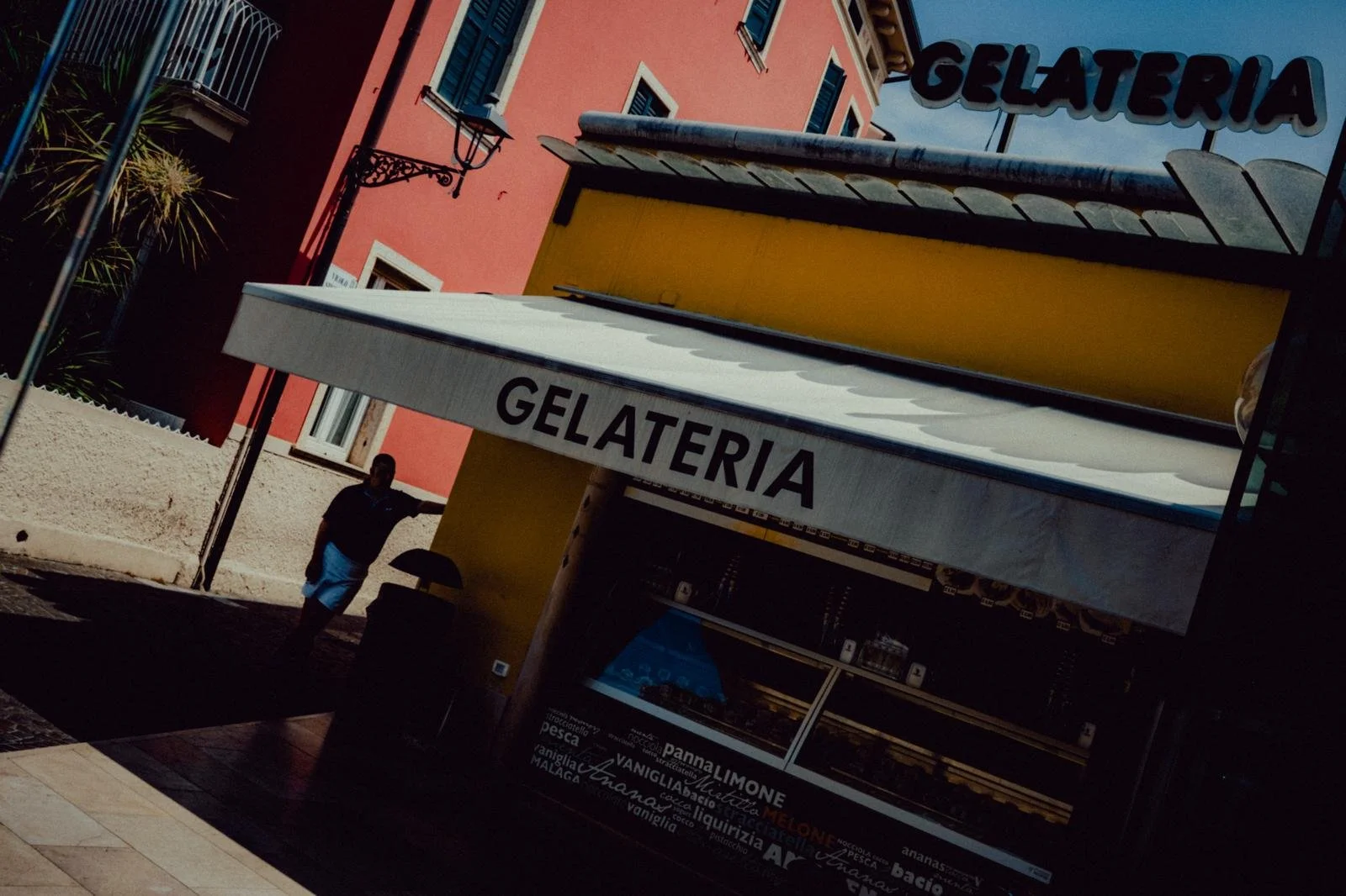 A gelateria shop with a white awning displaying the word 'GELATERIA' in black, and a counter inside visible behind glass showcasing ice cream flavors, set in an outdoor area with yellow and pink buildings in the background. A person in dark clothing