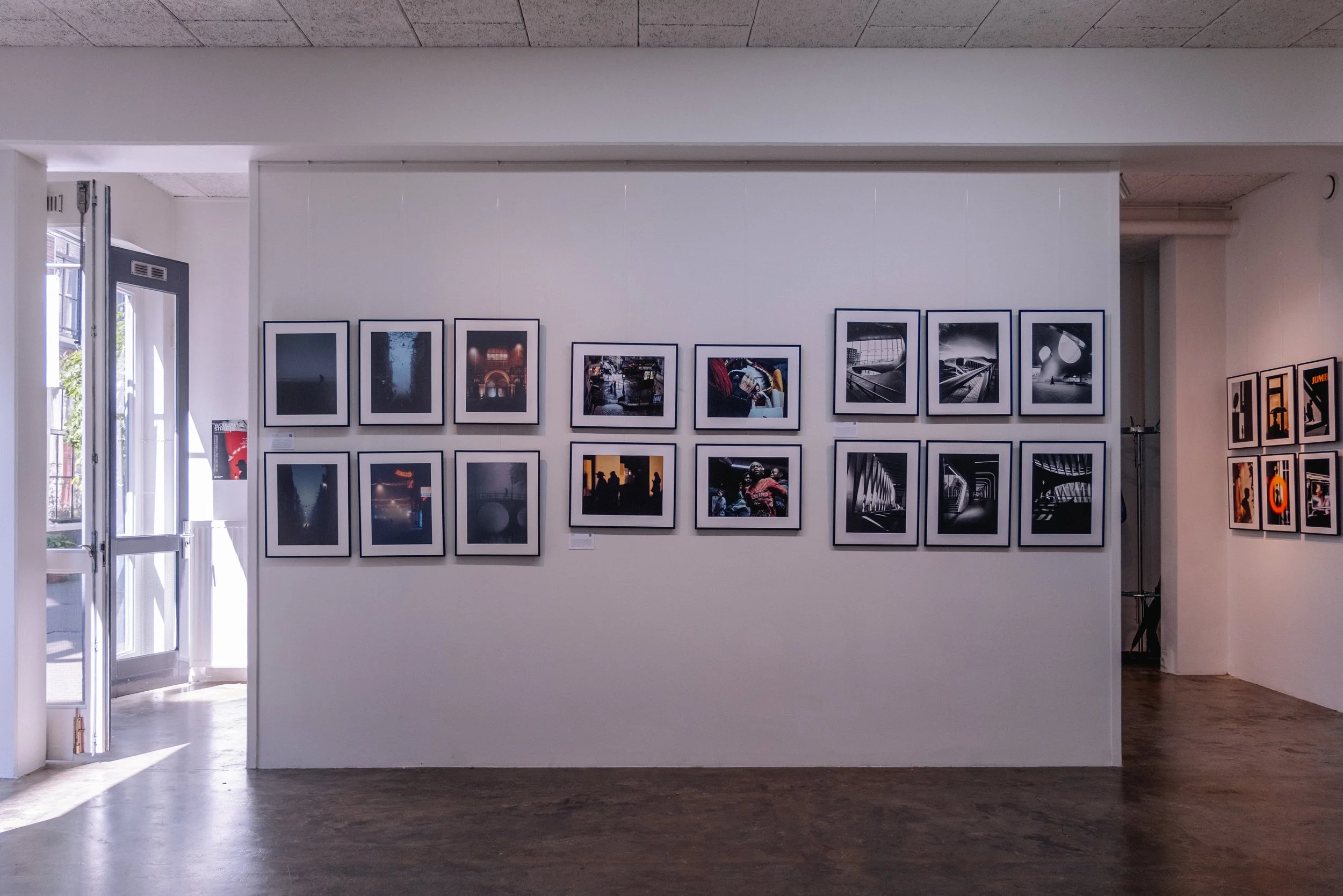 Inside an art gallery with framed photographs on white walls, natural light coming through an open door, and hardwood floors.
